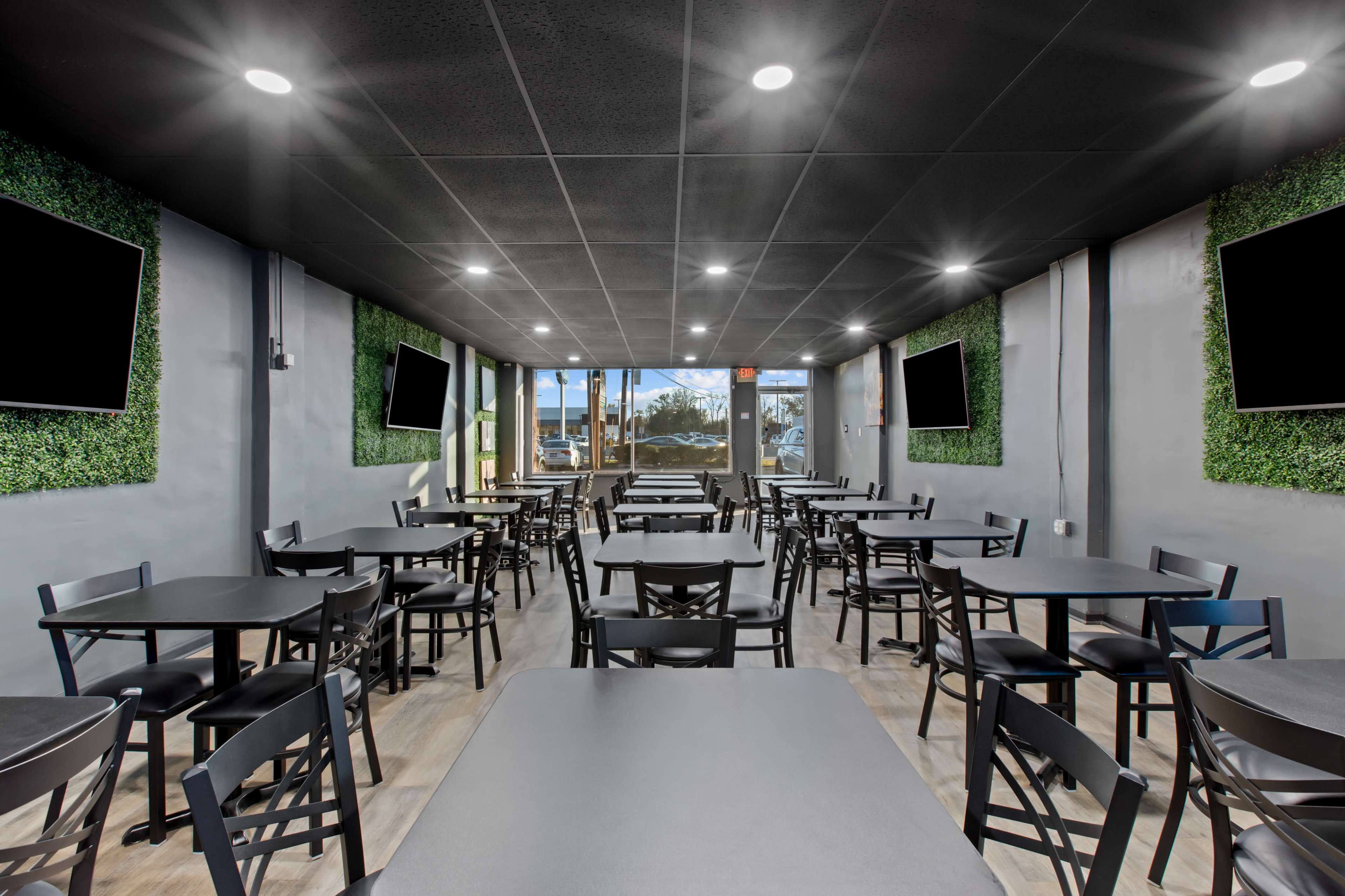 A modern dining area with multiple dark tables and chairs arranged in rows, flanked by green wall accents and large windows at one end.