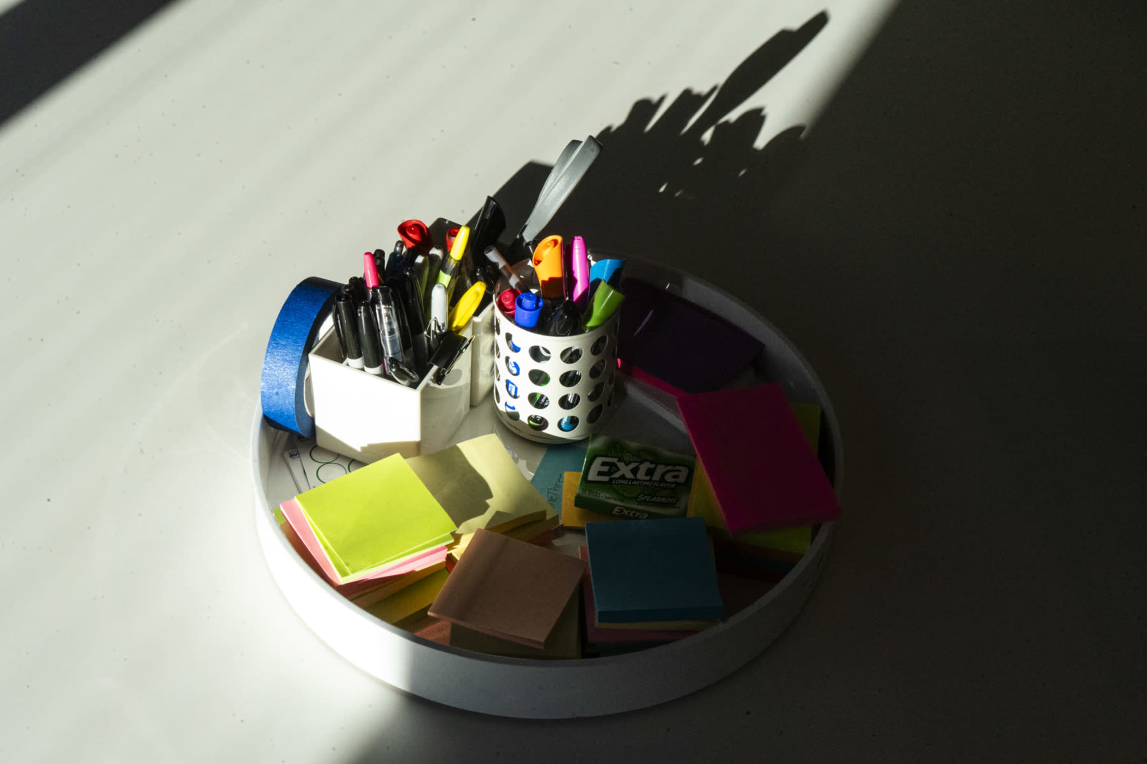 A round tray contains various colorful sticky notes and cups filled with pens and markers, illuminated by natural light creating shadows on the surface.