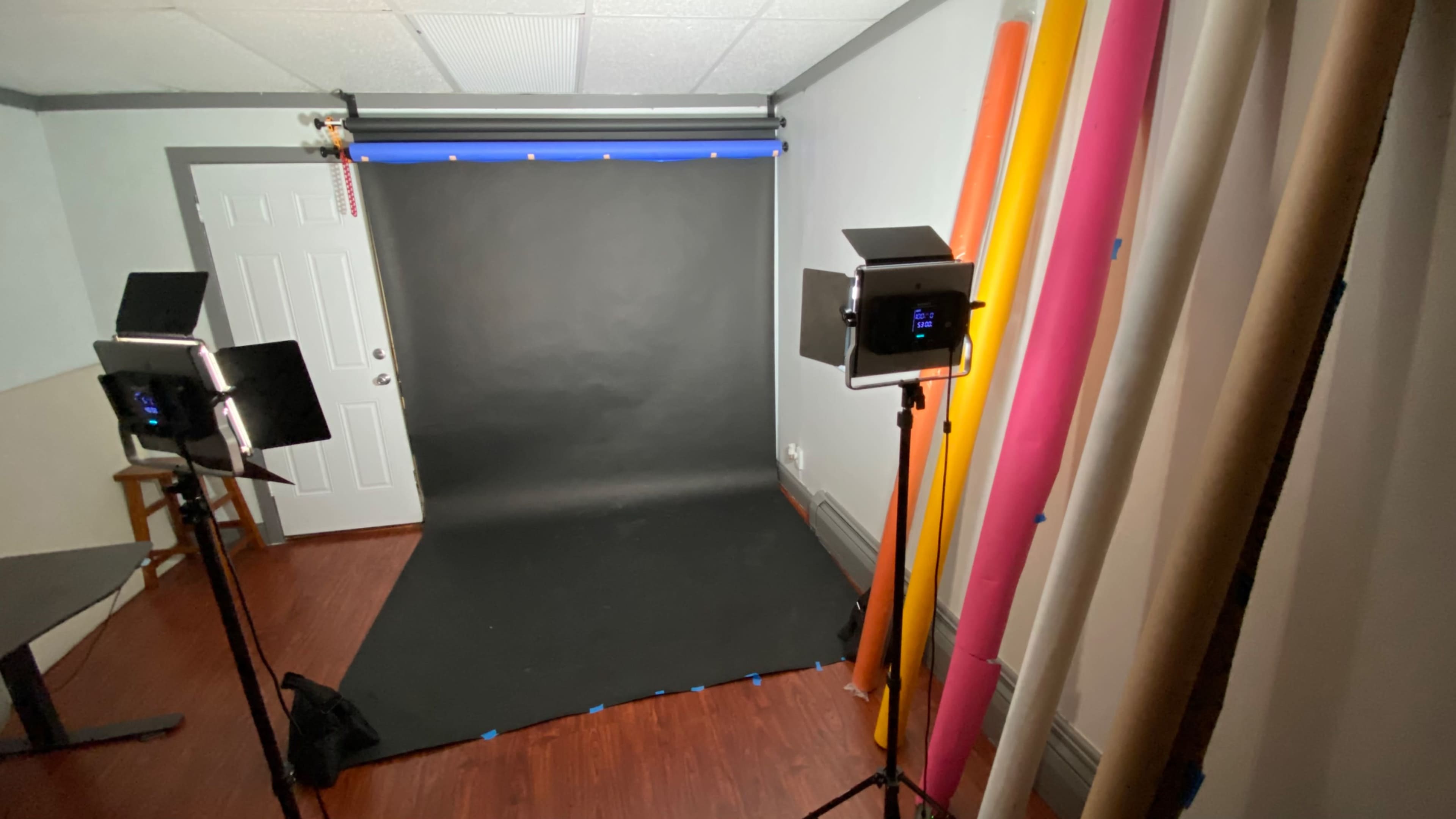 A photography studio with a black backdrop, lighting equipment, and colorful rolls of paper on the sides.