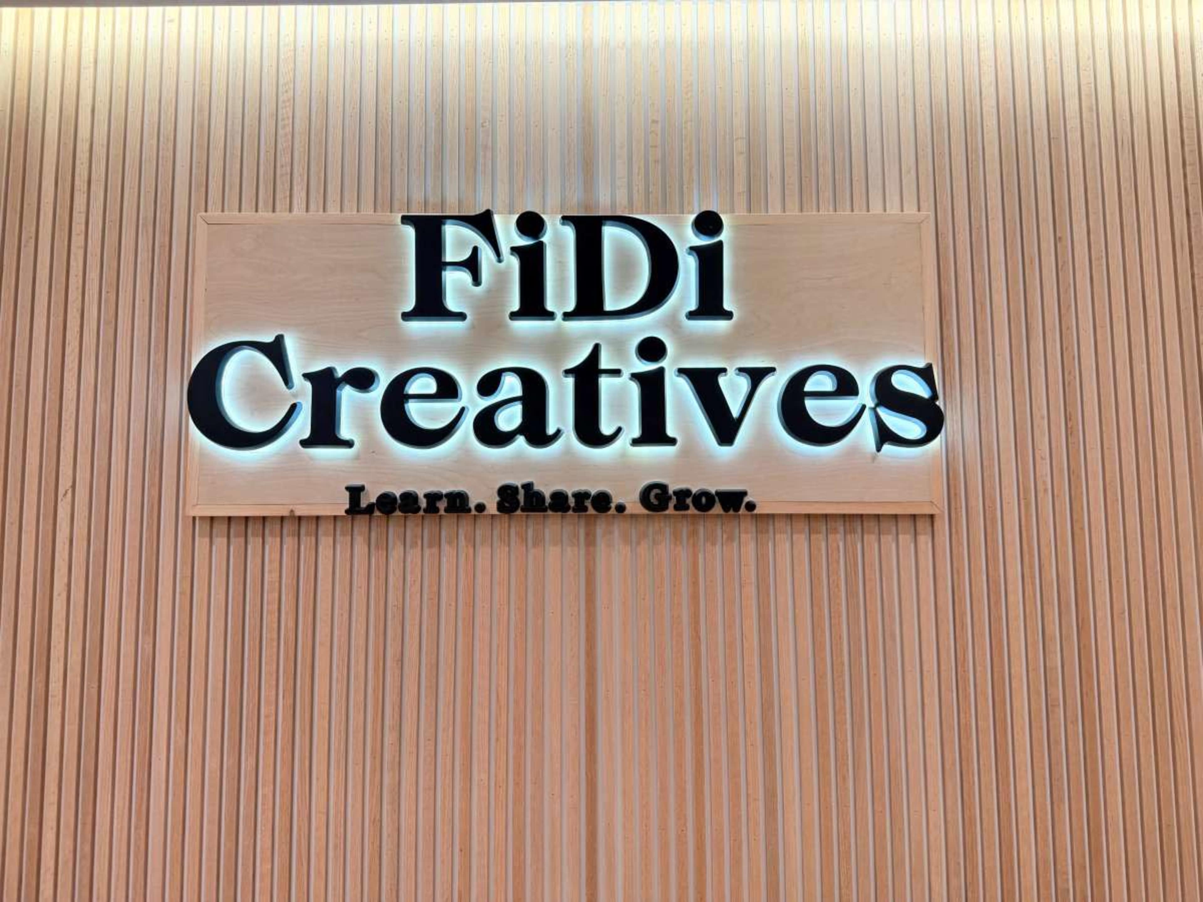 The image shows a wooden wall with a sign that reads "FiDi Creatives" in bold letters, accompanied by the tagline "Learn. Share. Grow."