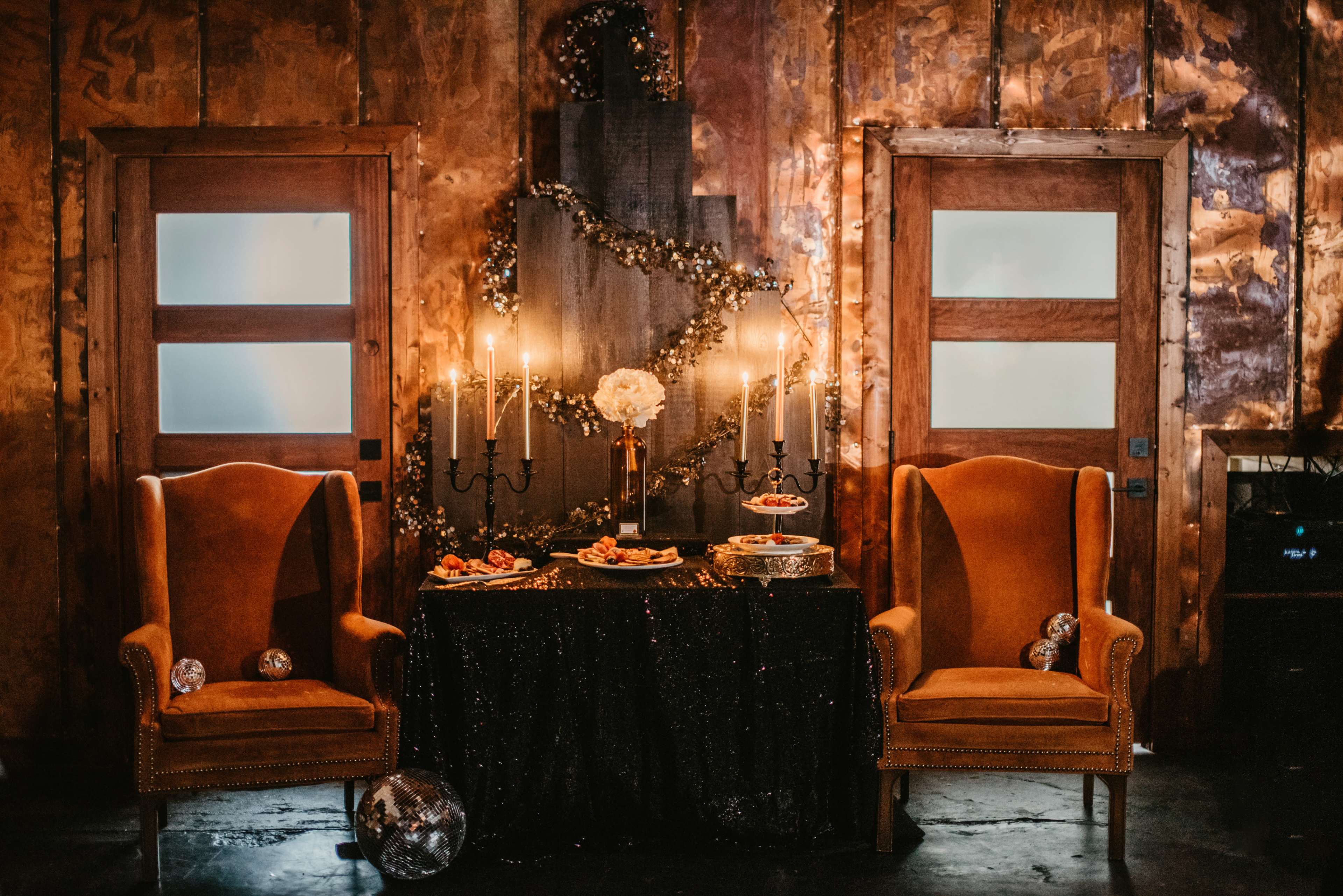 The scene features two orange armchairs flanking a black table adorned with a cake and snacks, set against a backdrop of wooden walls decorated with garlands and candles.