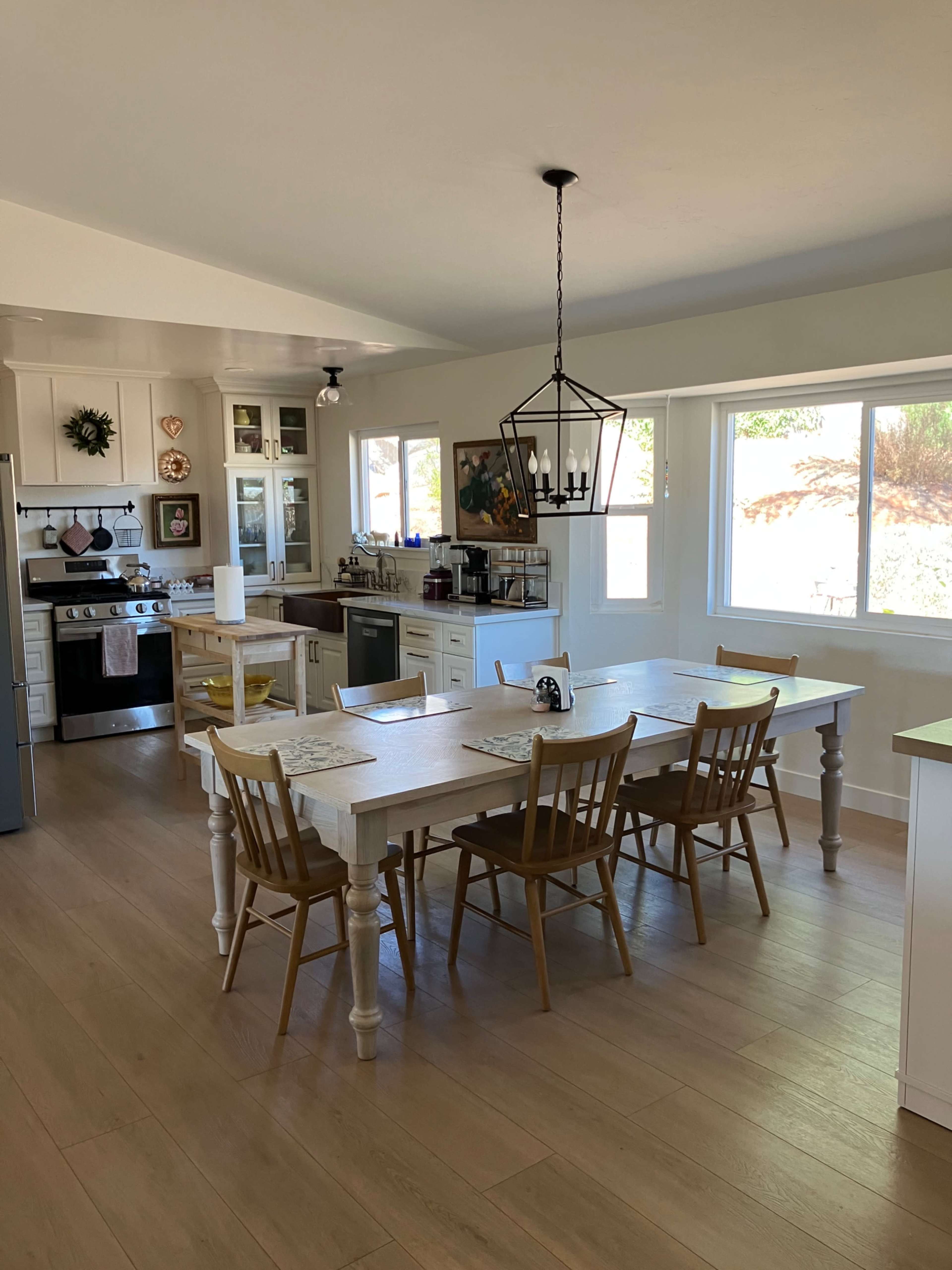 The image shows a spacious kitchen with a dining table, six chairs, and large windows allowing natural light.