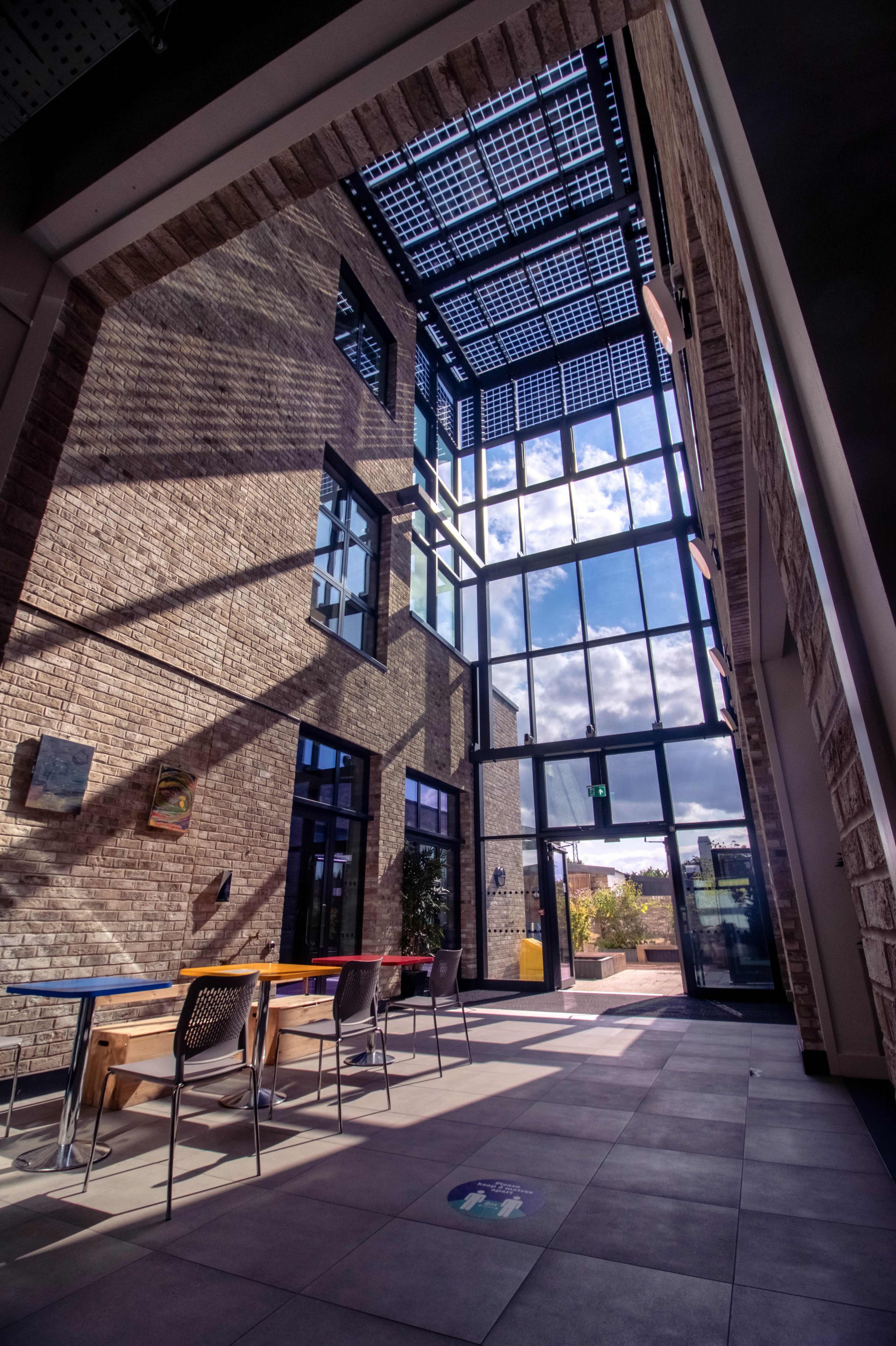 A modern architectural space with large windows, exposed brick walls, and a series of tables and chairs arranged along the floor.
