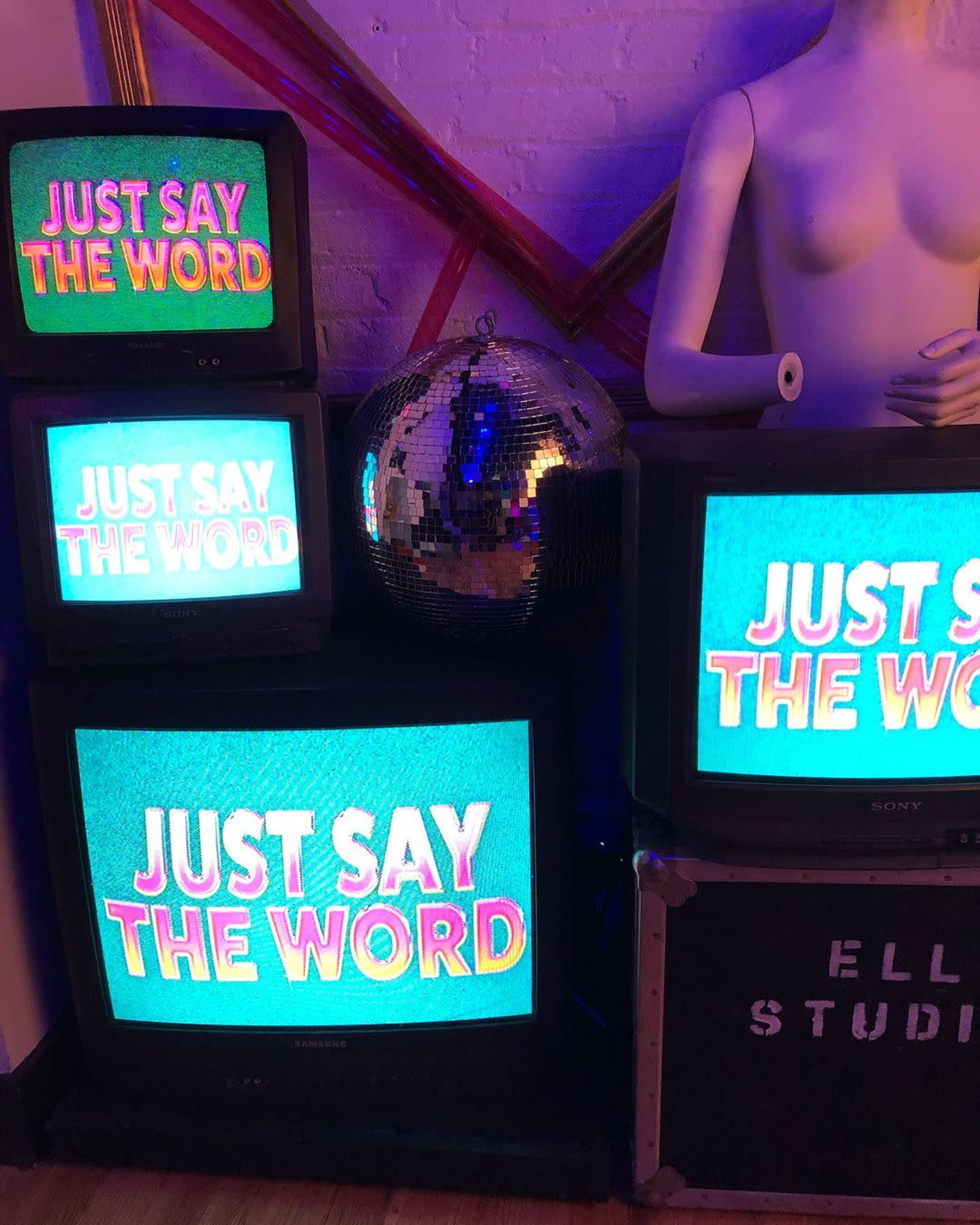 The image features multiple television sets stacked together, displaying the text "JUST SAY THE WORD" in bright colors, alongside a reflective disco ball and a mannequin torso.