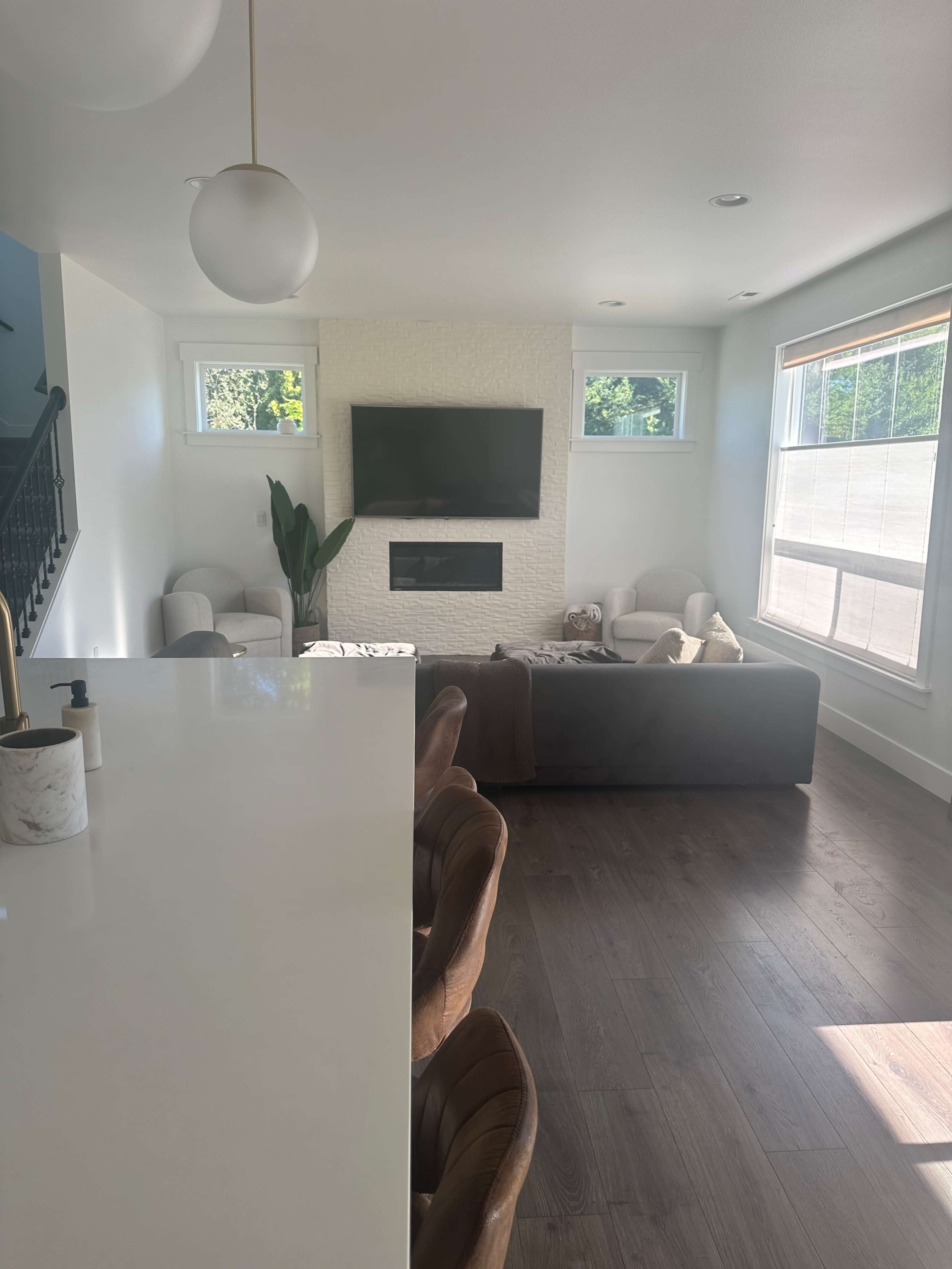 A modern living room with a television mounted above a white stone fireplace, complemented by two armchairs and a sofa, all bathed in natural light from large windows.