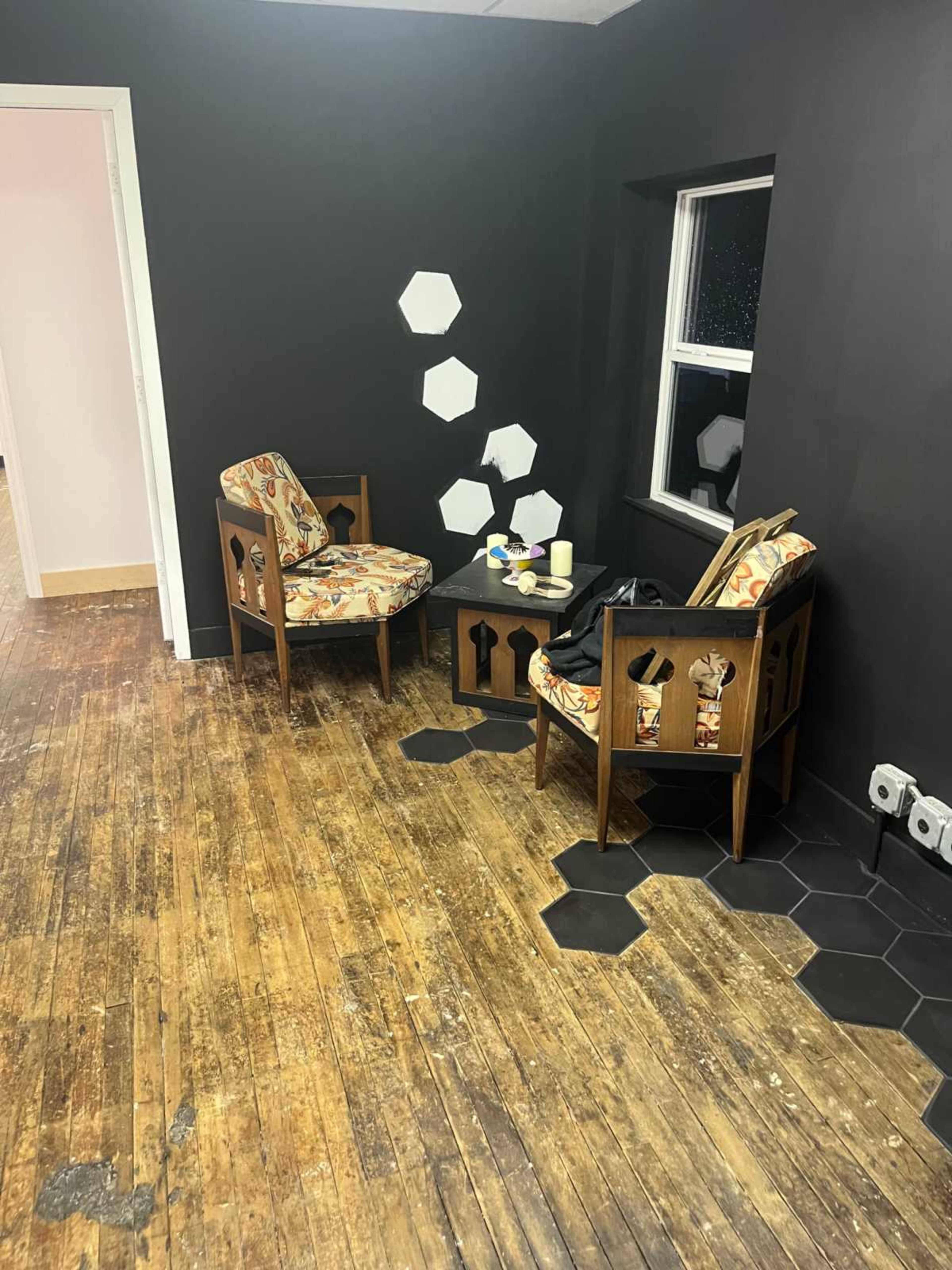 The image shows a small corner of a room featuring two patterned armchairs, a small table, and a black hexagonal floor motif against a dark wall.