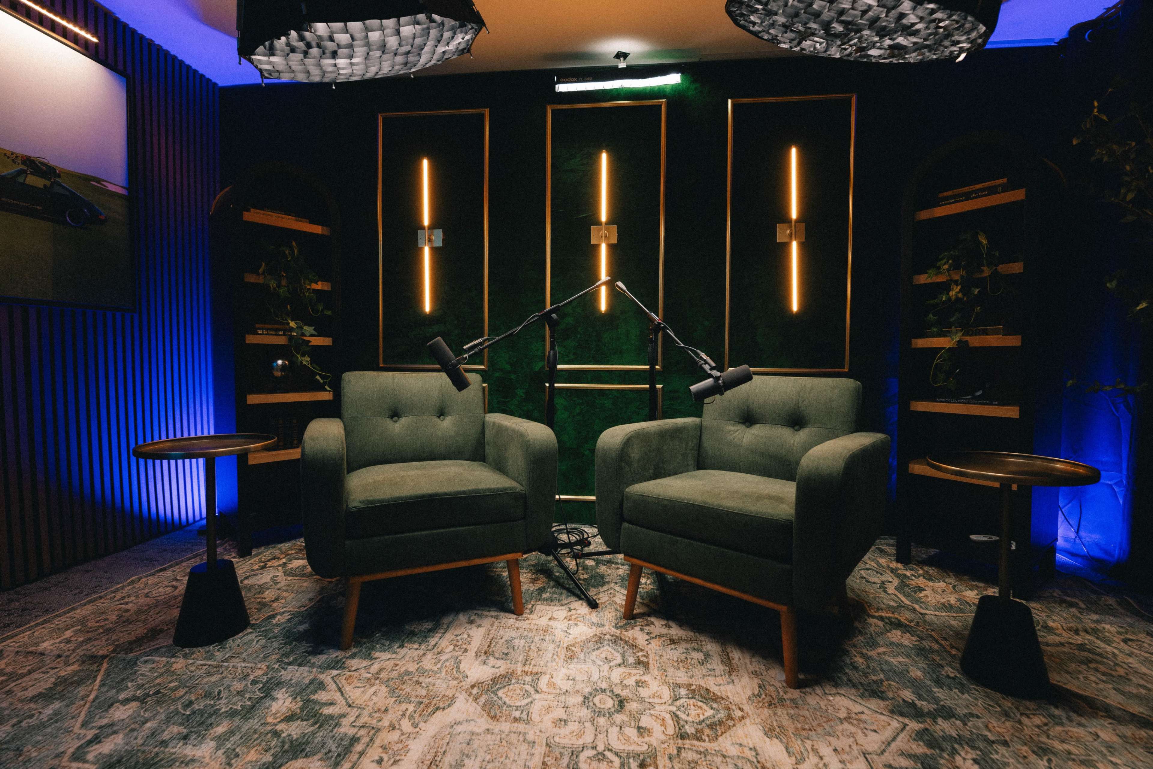 The image shows a stylish interview setup featuring two green armchairs, a patterned rug, and a backdrop with ambient lighting and shelves.