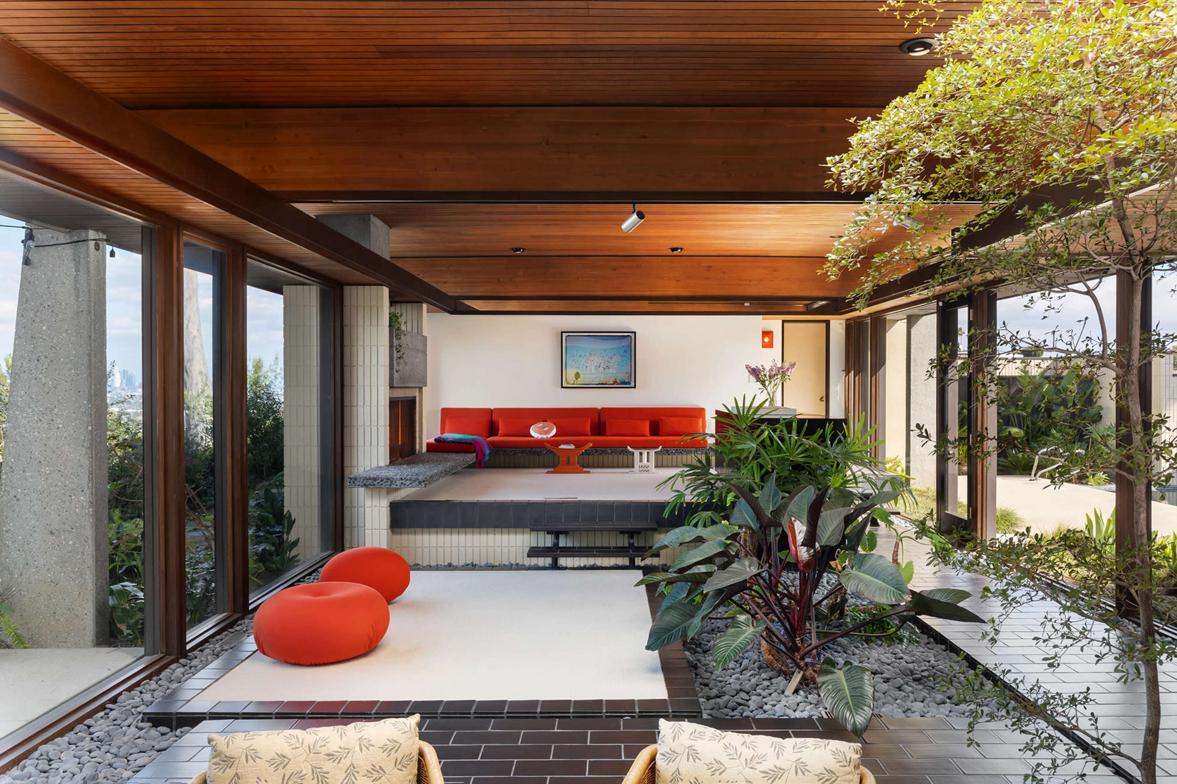 The image shows a modern interior space featuring a sunken seating area with an orange couch and two round cushions, surrounded by plants and large windows that provide a view of the outdoors.