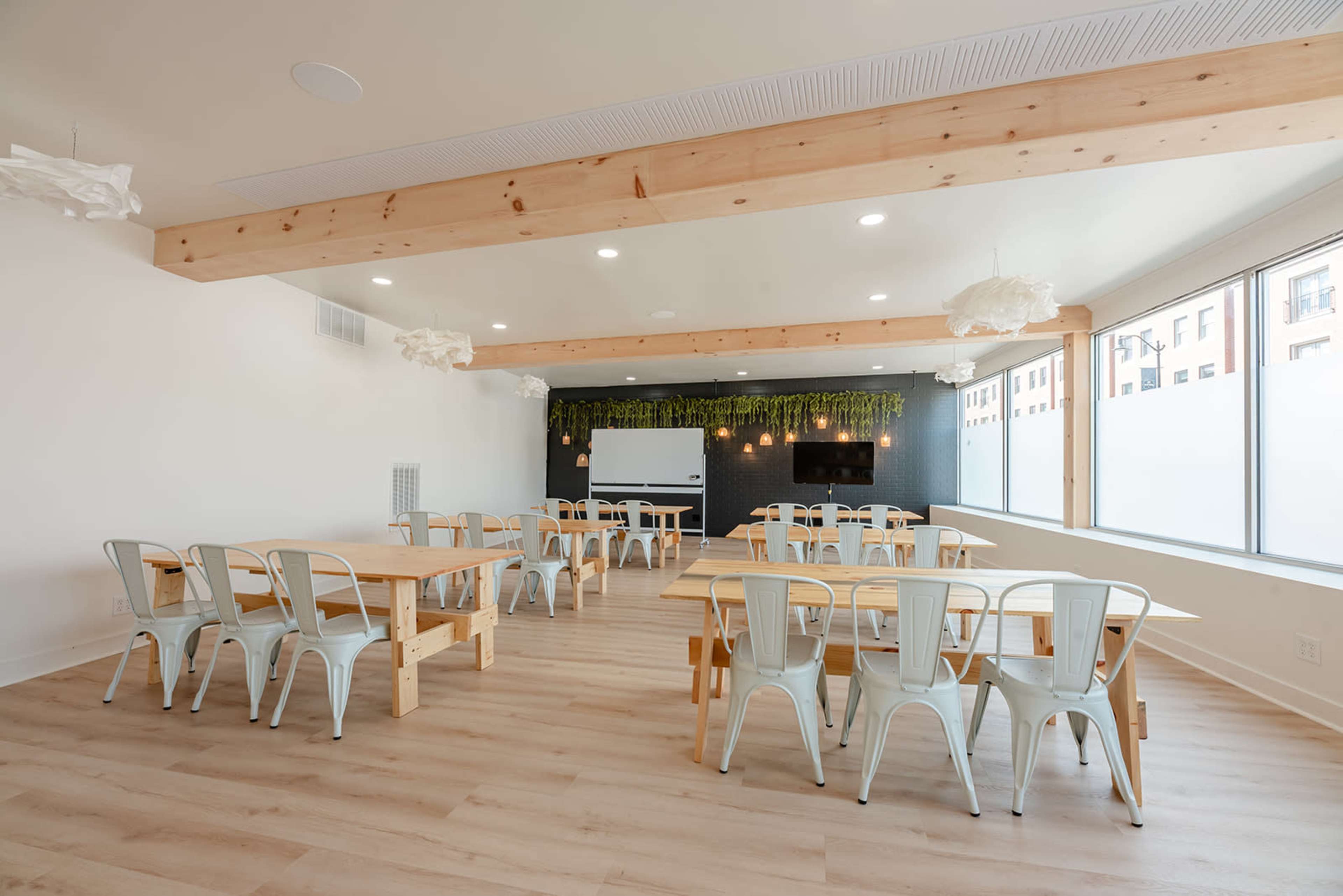 A spacious, modern classroom with wooden beams, multiple wooden tables, and metal chairs arranged in a bright, well-lit environment.