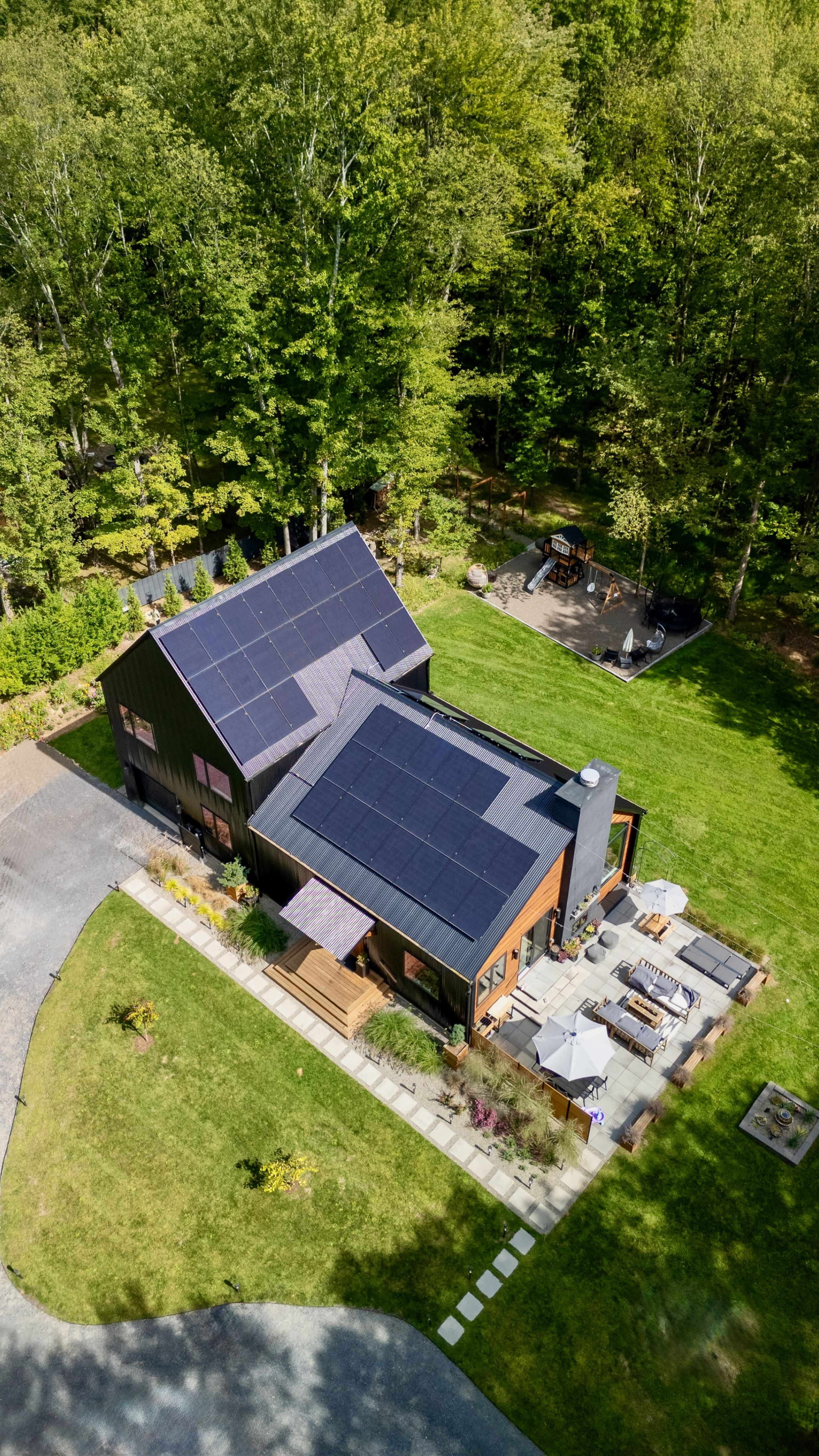 The image shows a modern house with solar panels on the roof, surrounded by trees and a well-maintained lawn, featuring a patio area with outdoor furniture.