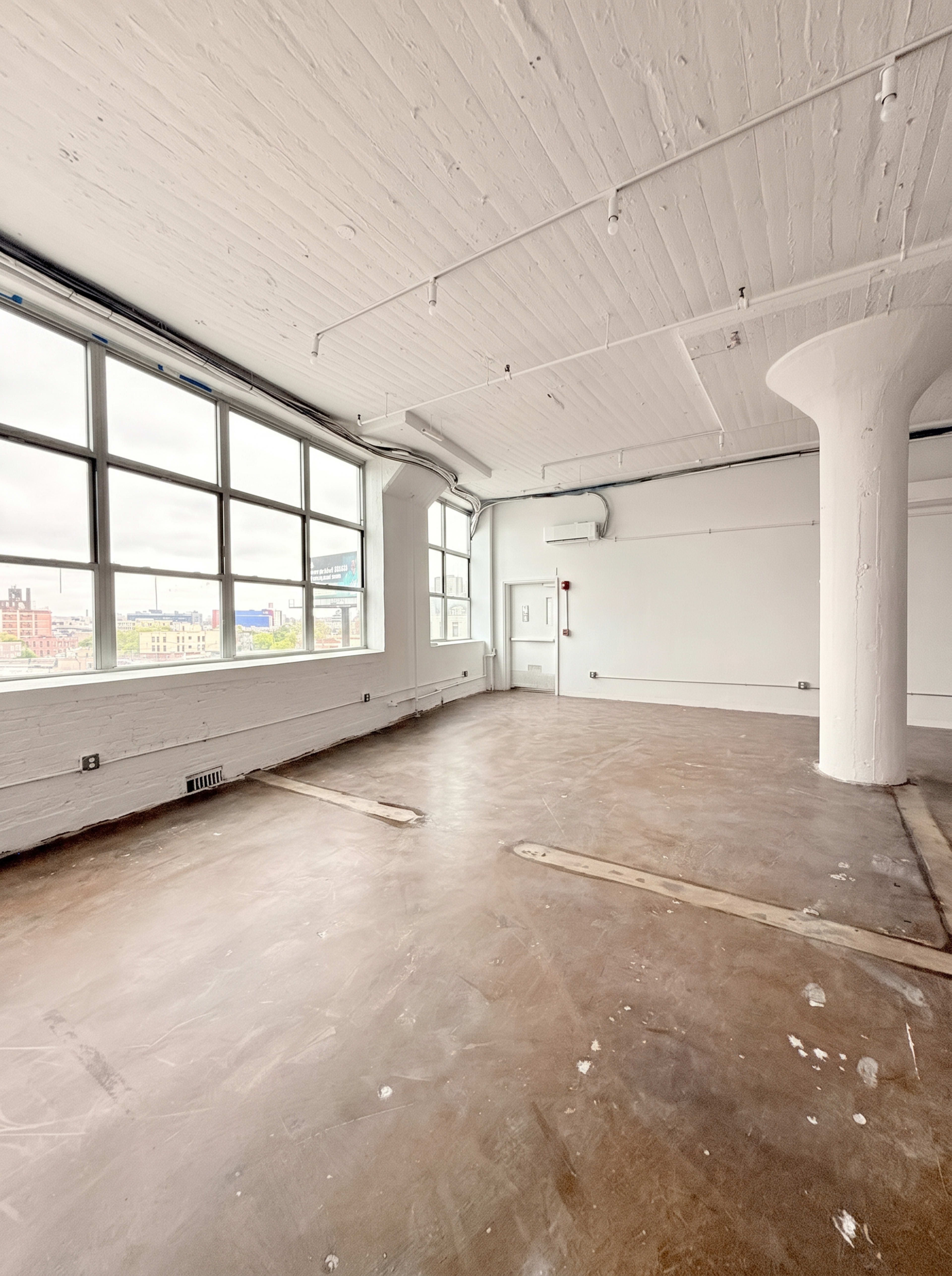 The image shows a spacious, empty room with large windows and exposed concrete walls and flooring.