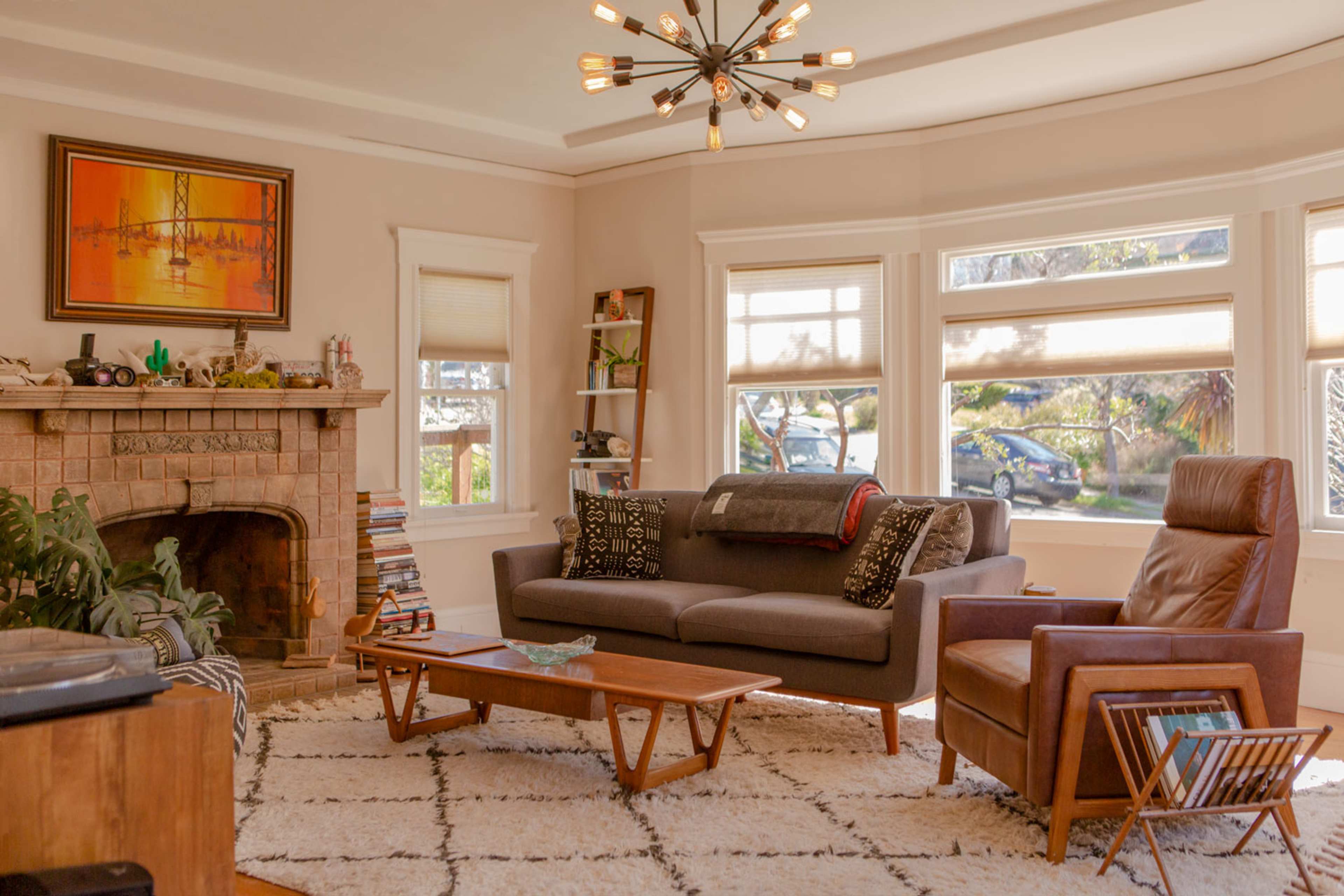 The image shows a cozy living room with a sectional sofa, a leather armchair, and a stone fireplace, complemented by large windows that let in natural light.