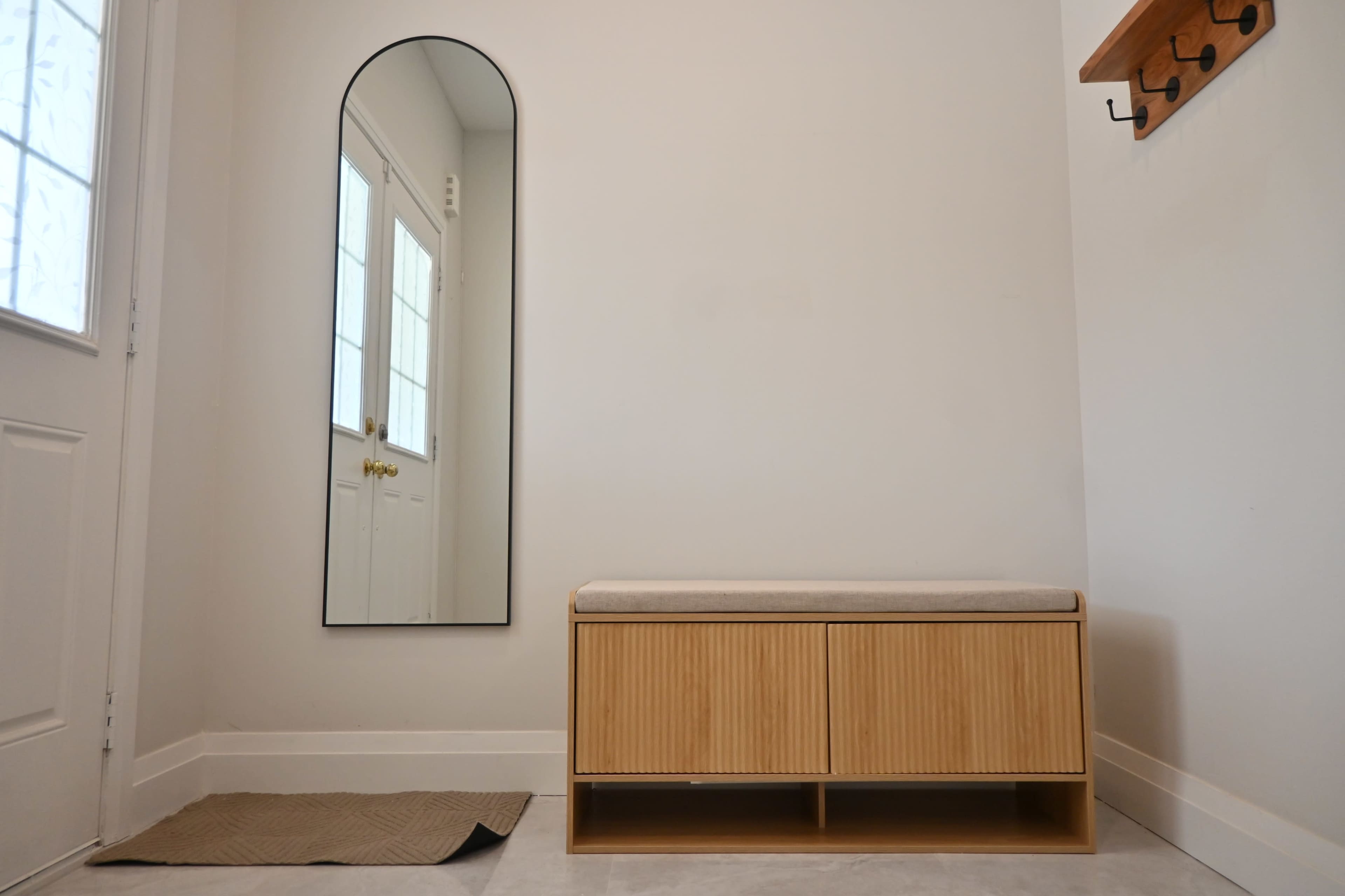 The image shows a minimalist entryway featuring a wooden storage bench with a cushioned top, a tall mirror, and a wall hook rack near the front door.