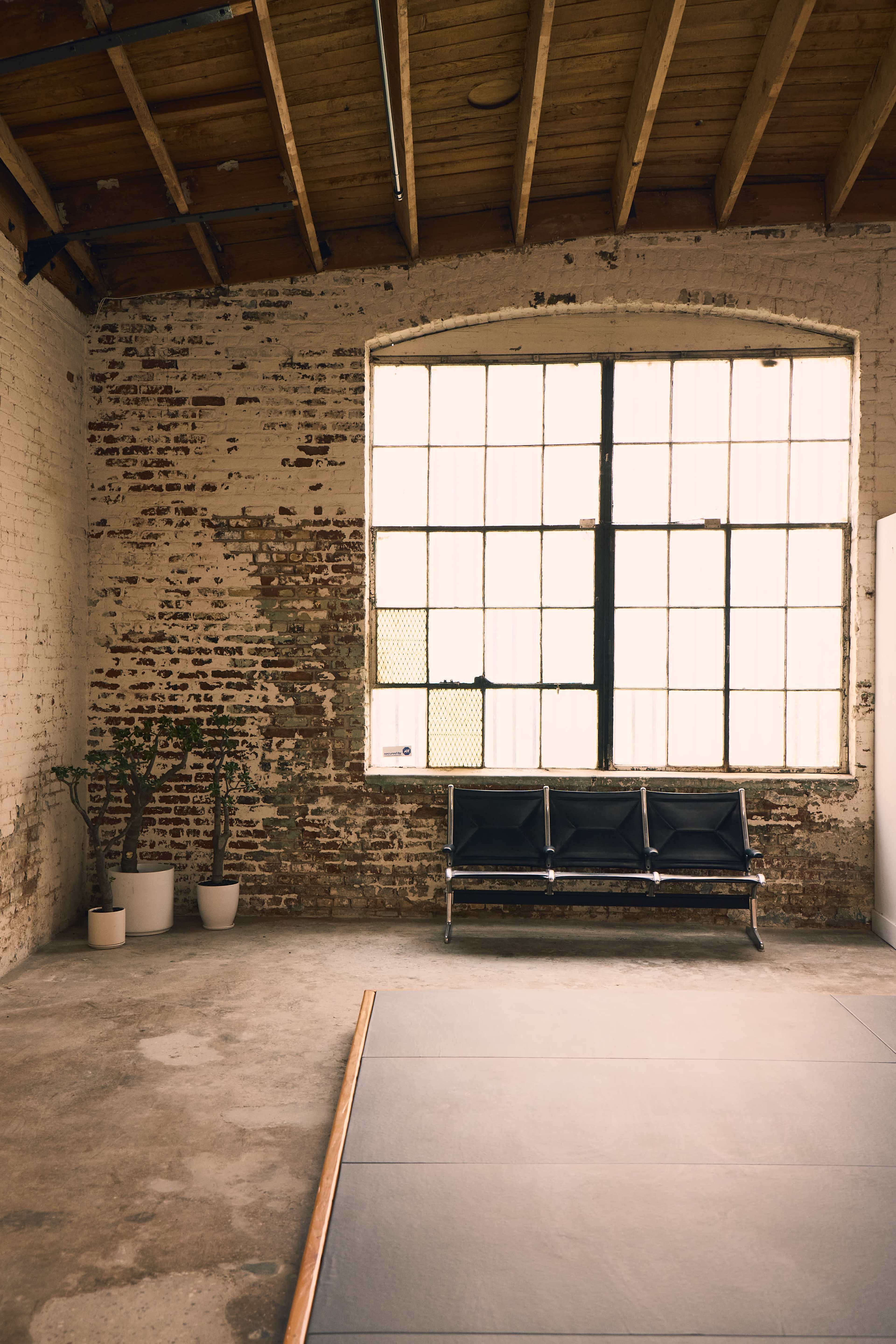 The image shows an industrial-style room with exposed brick walls, a large window, and a minimalist seating area featuring a black bench.