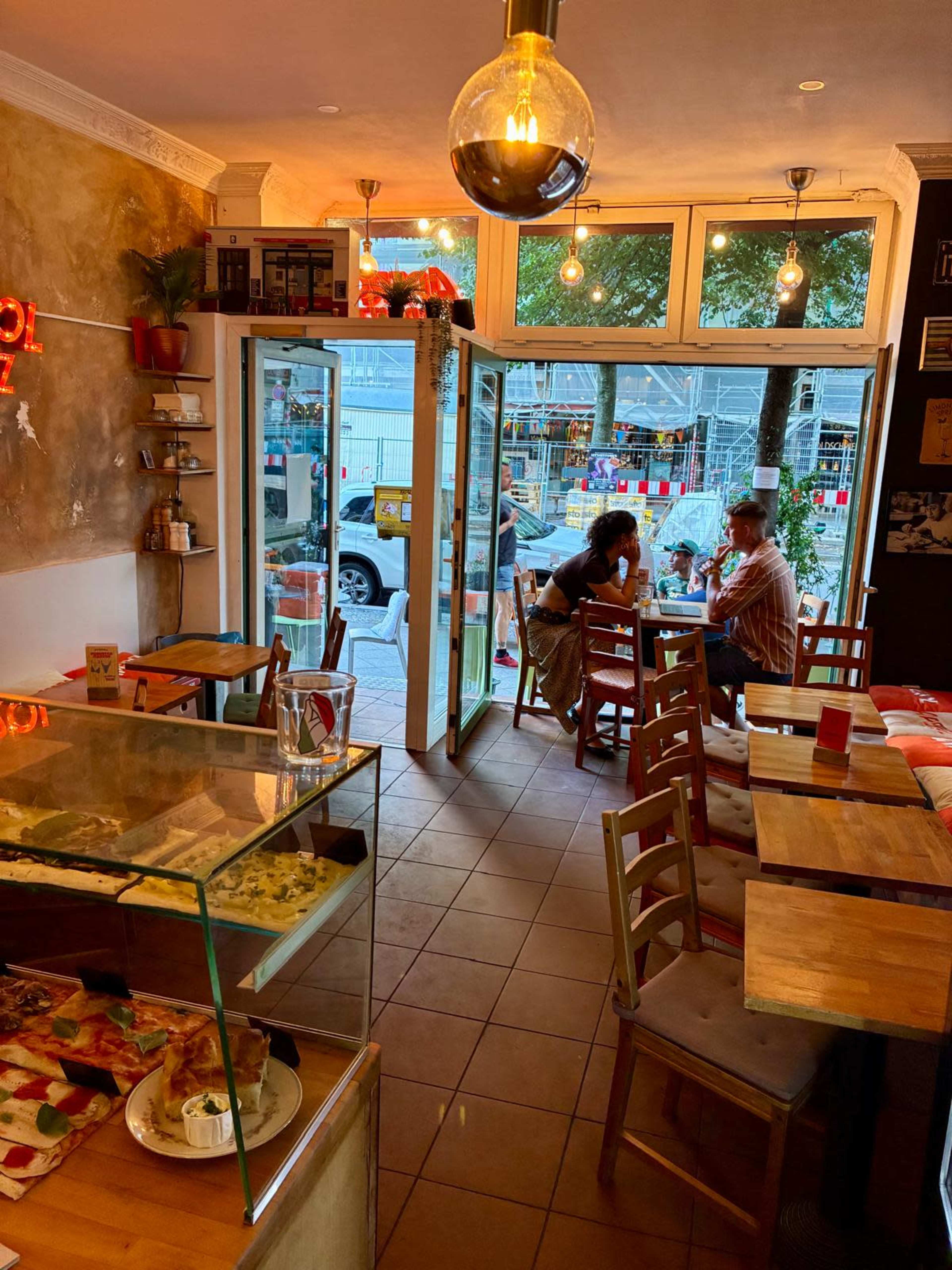 The image shows an interior view of a cozy café with wooden tables, a display case of food, and customers seated at tables near glass doors that open to the street.