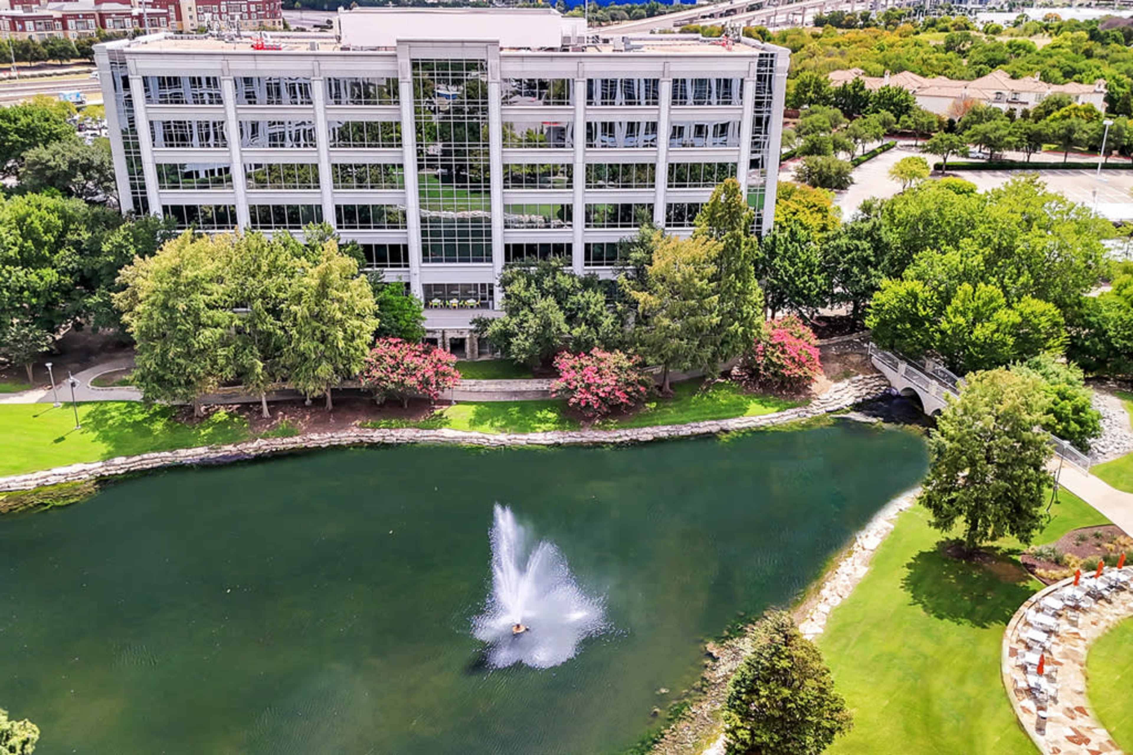 A modern building overlooks a tranquil pond with a fountain and lush greenery surrounding it.