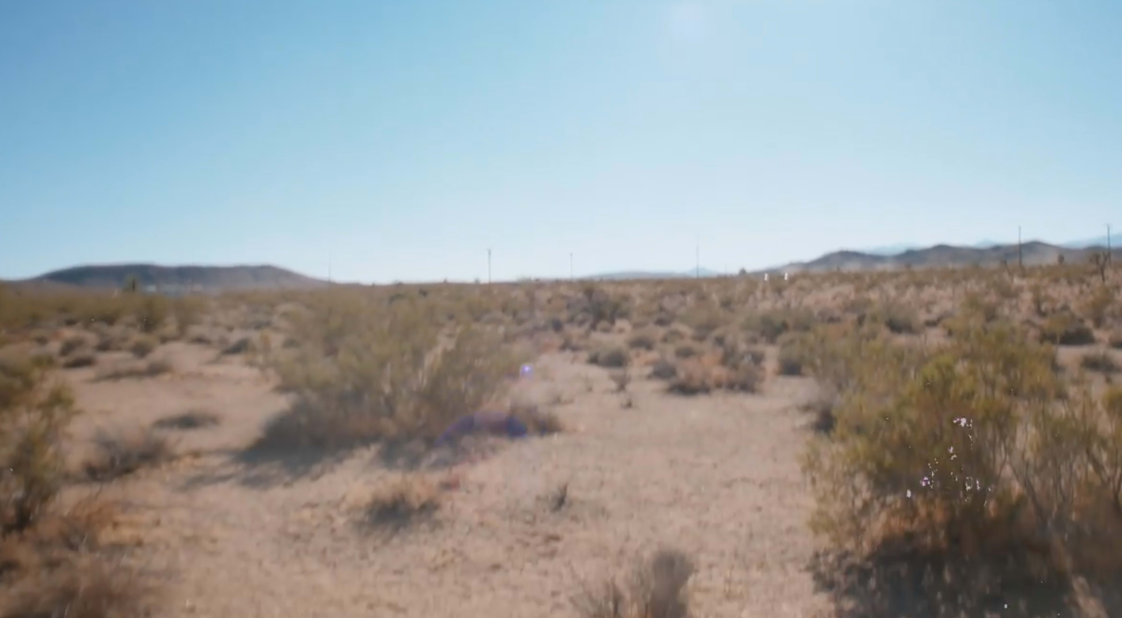 The image shows a wide, arid landscape with sparse vegetation and distant hills under a clear blue sky.