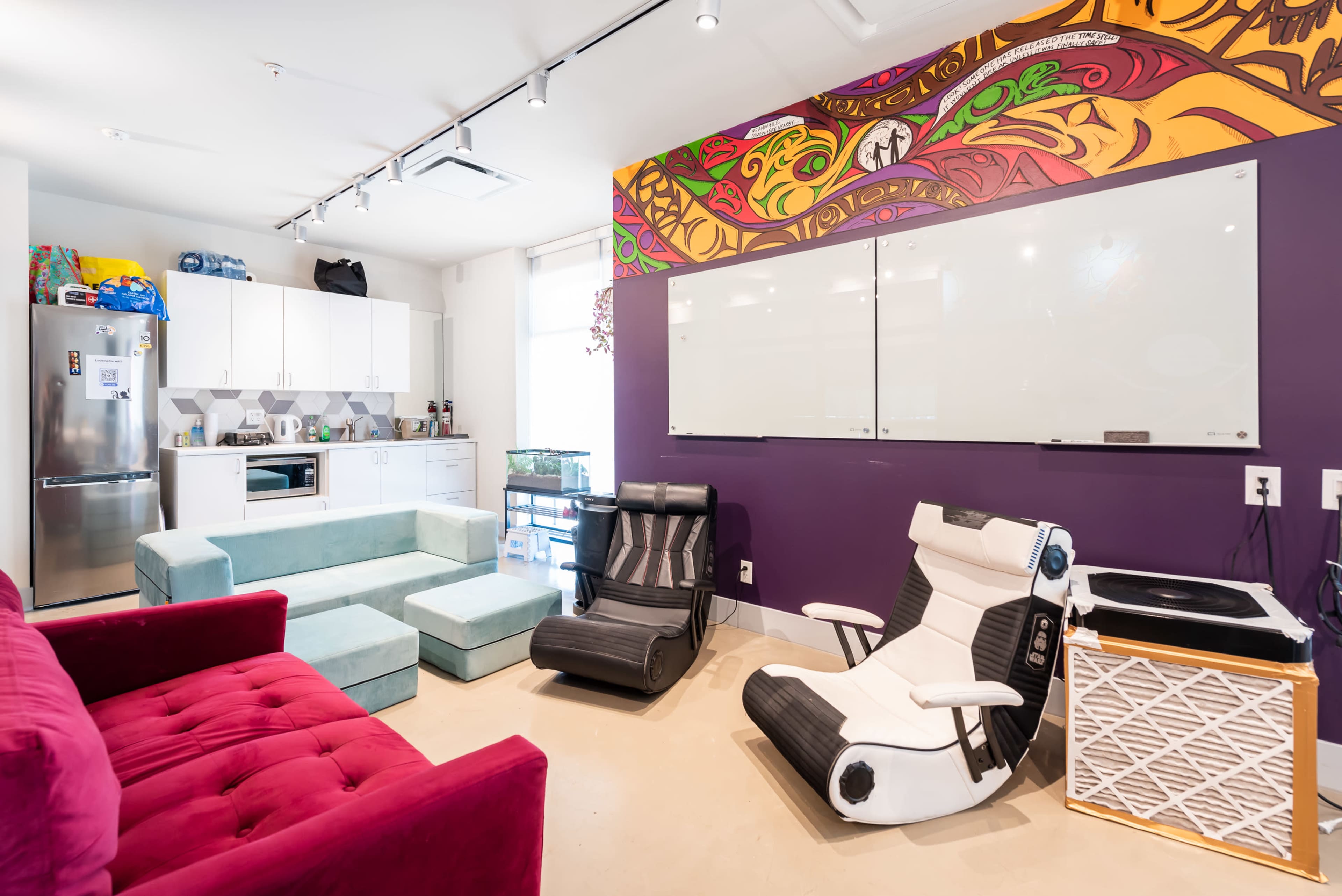 A modern lounge area with colorful walls, seating options including a red velvet couch and two lounge chairs, and a kitchenette in the background.