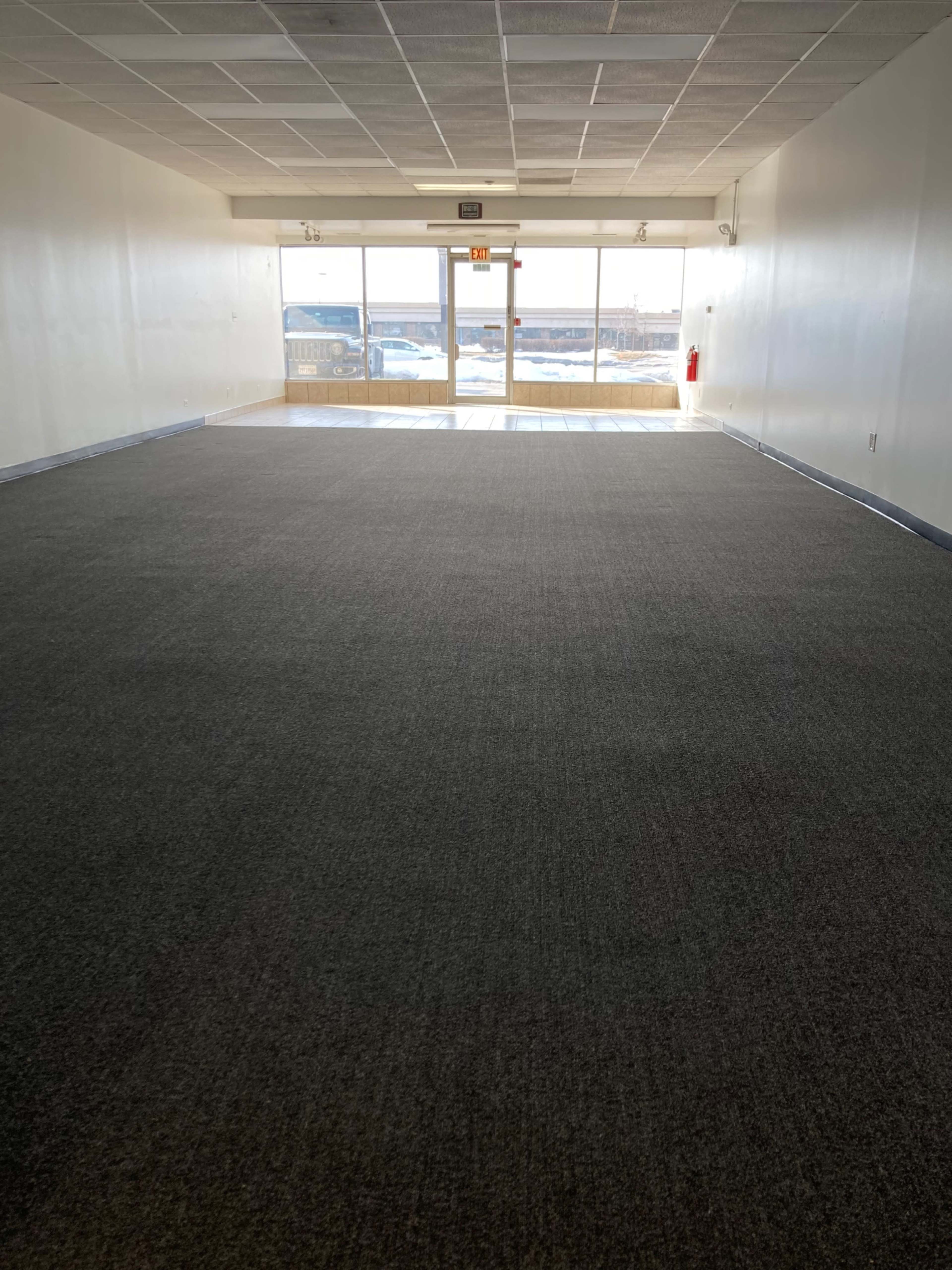 A large, empty room with a carpeted floor and a wide glass entrance at the far end.