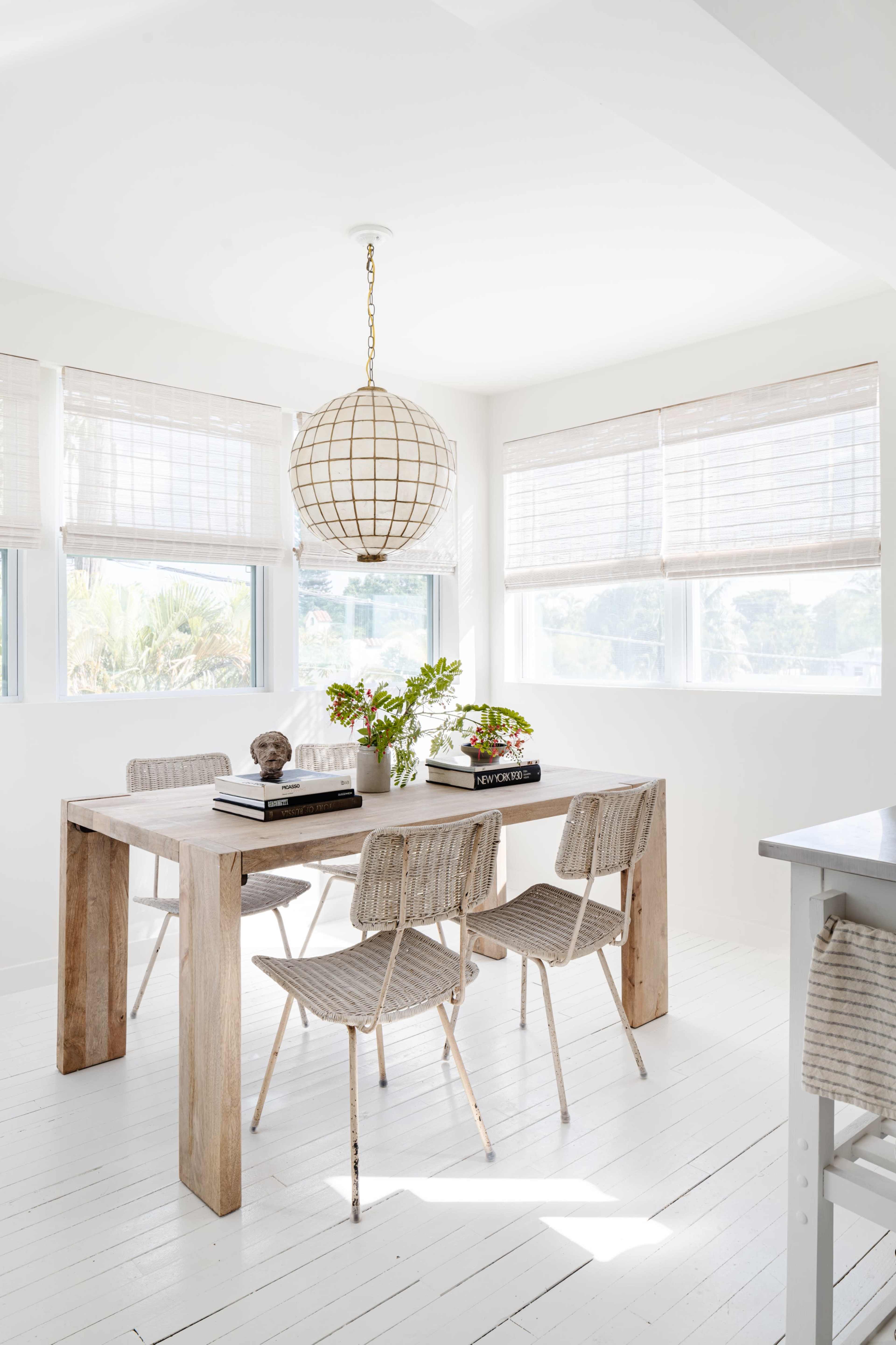 A bright dining area features a wooden table surrounded by woven chairs, with a large pendant light overhead and windows dressed in light blinds.