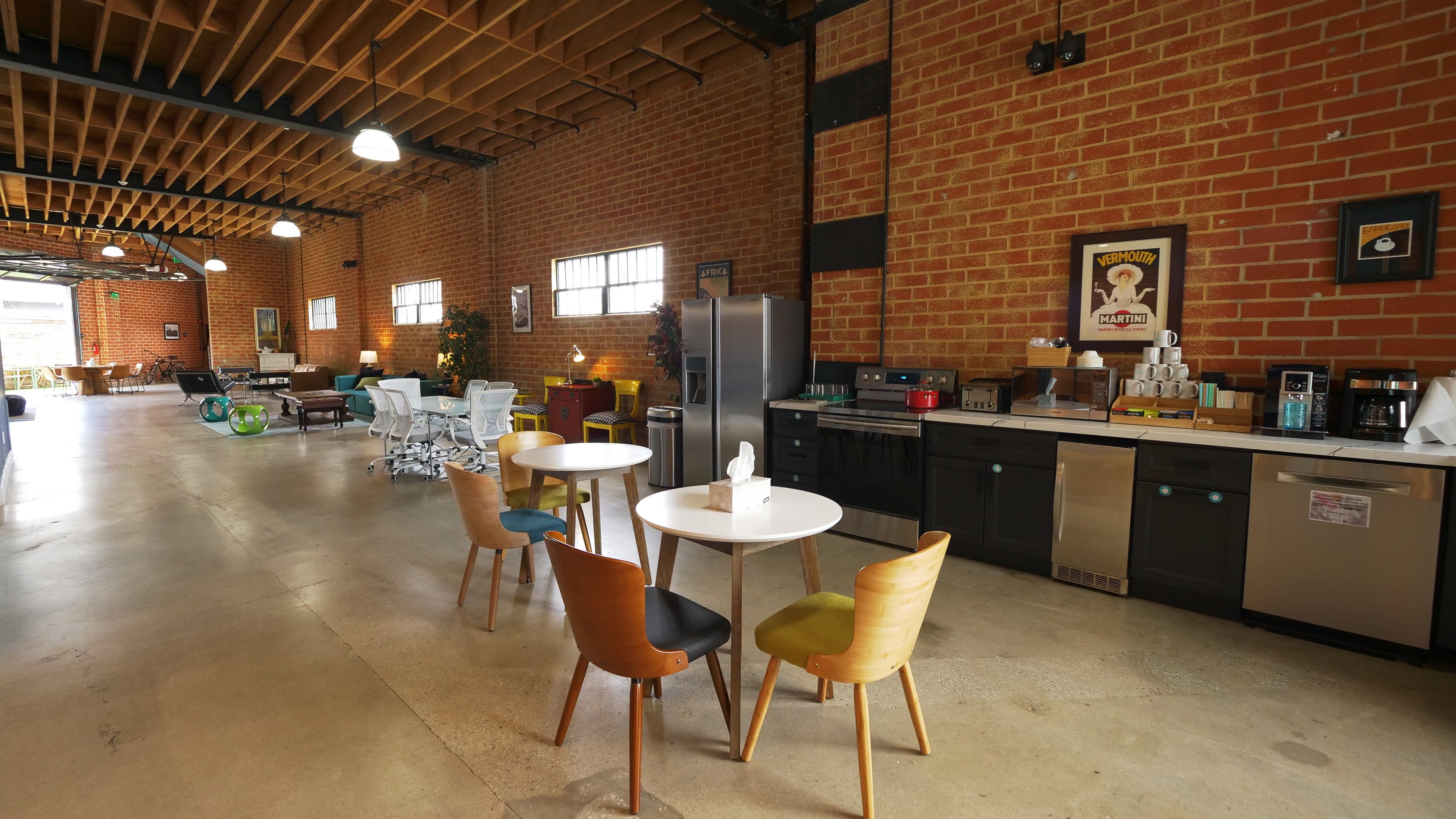 The image shows a spacious industrial-style kitchen with a dining area featuring colorful chairs around a white table, alongside modern appliances and brick walls.