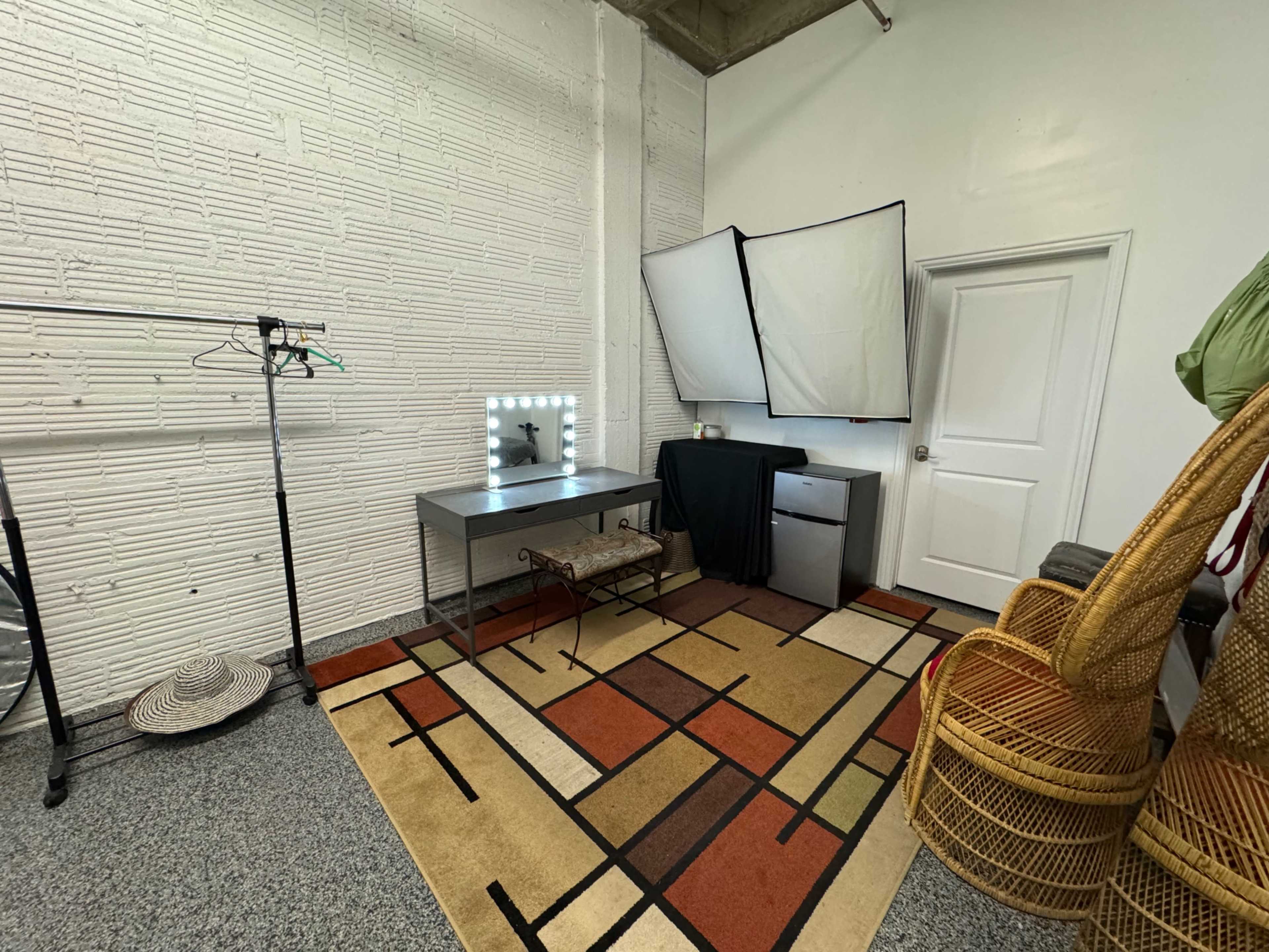A small studio space featuring a vanity mirror with lights, a desk, a rolling clothing rack, two wicker chairs, and a backdrop setup, all arranged on a patterned rug.