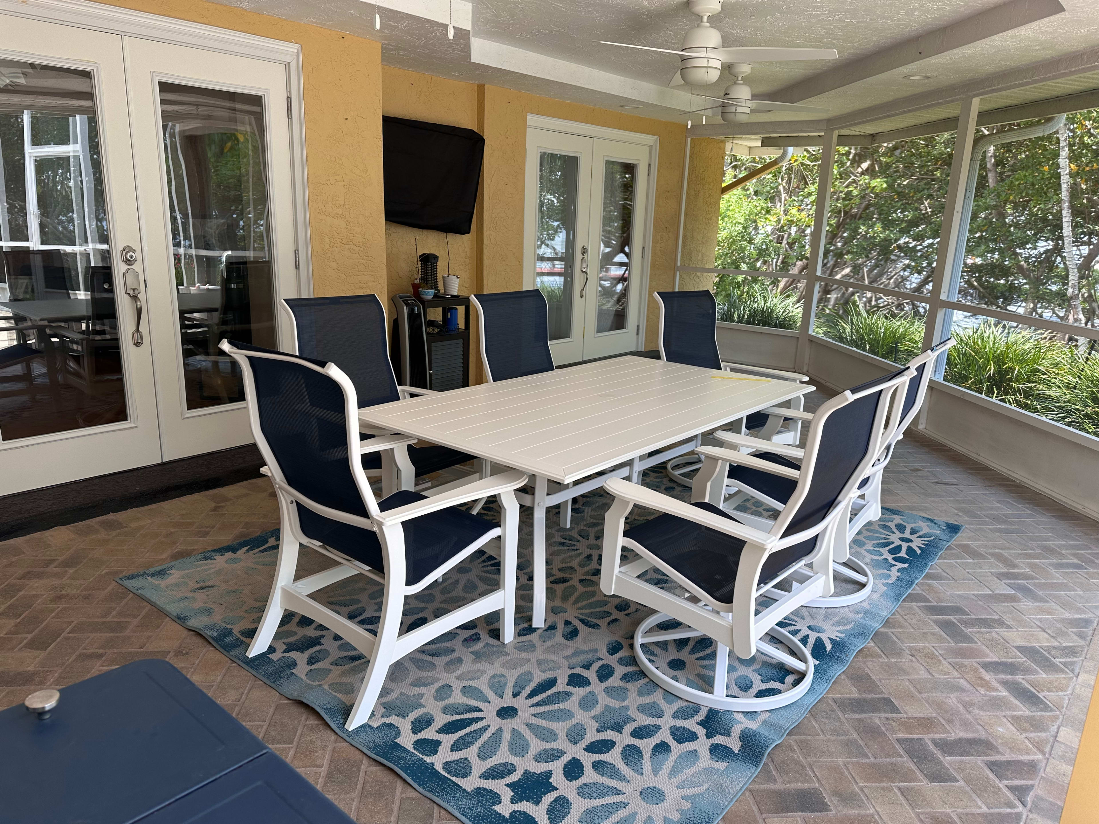 A screened porch features a white patio table surrounded by six navy blue chairs, with a patterned rug underneath and sliding glass doors in the background.