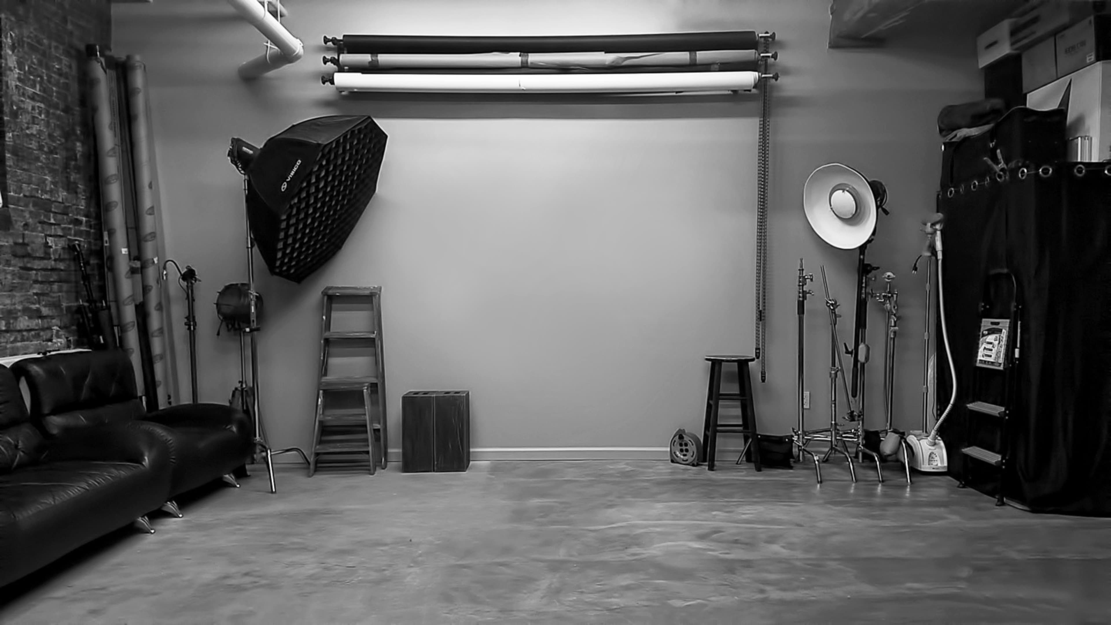 The image shows a minimalist photography studio with various lighting equipment, a black sofa, and wooden furniture against a plain wall.