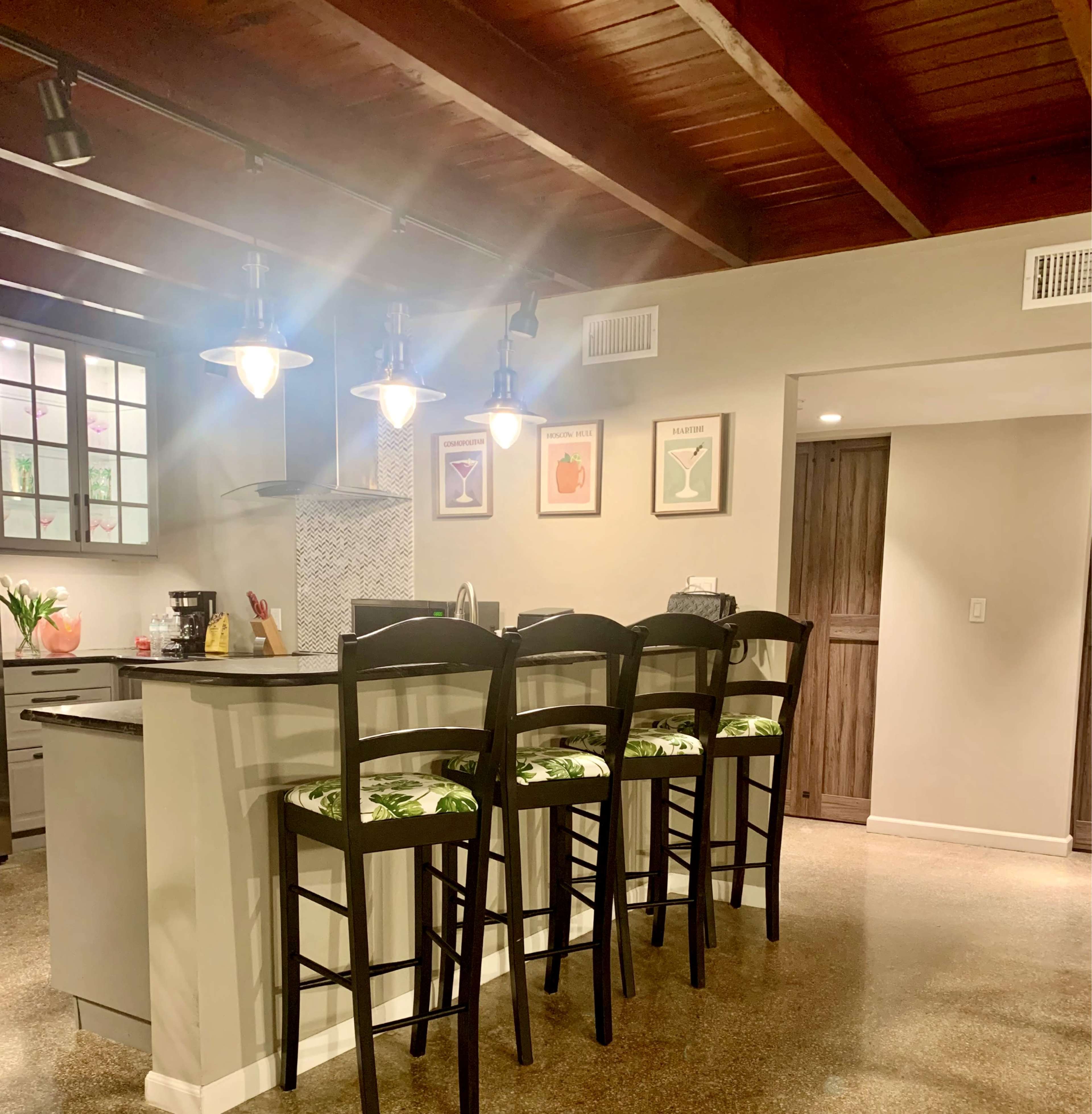 The image depicts a modern kitchen with a bar area featuring four tall chairs, a countertop, and decorative fruit-themed artwork on the wall.