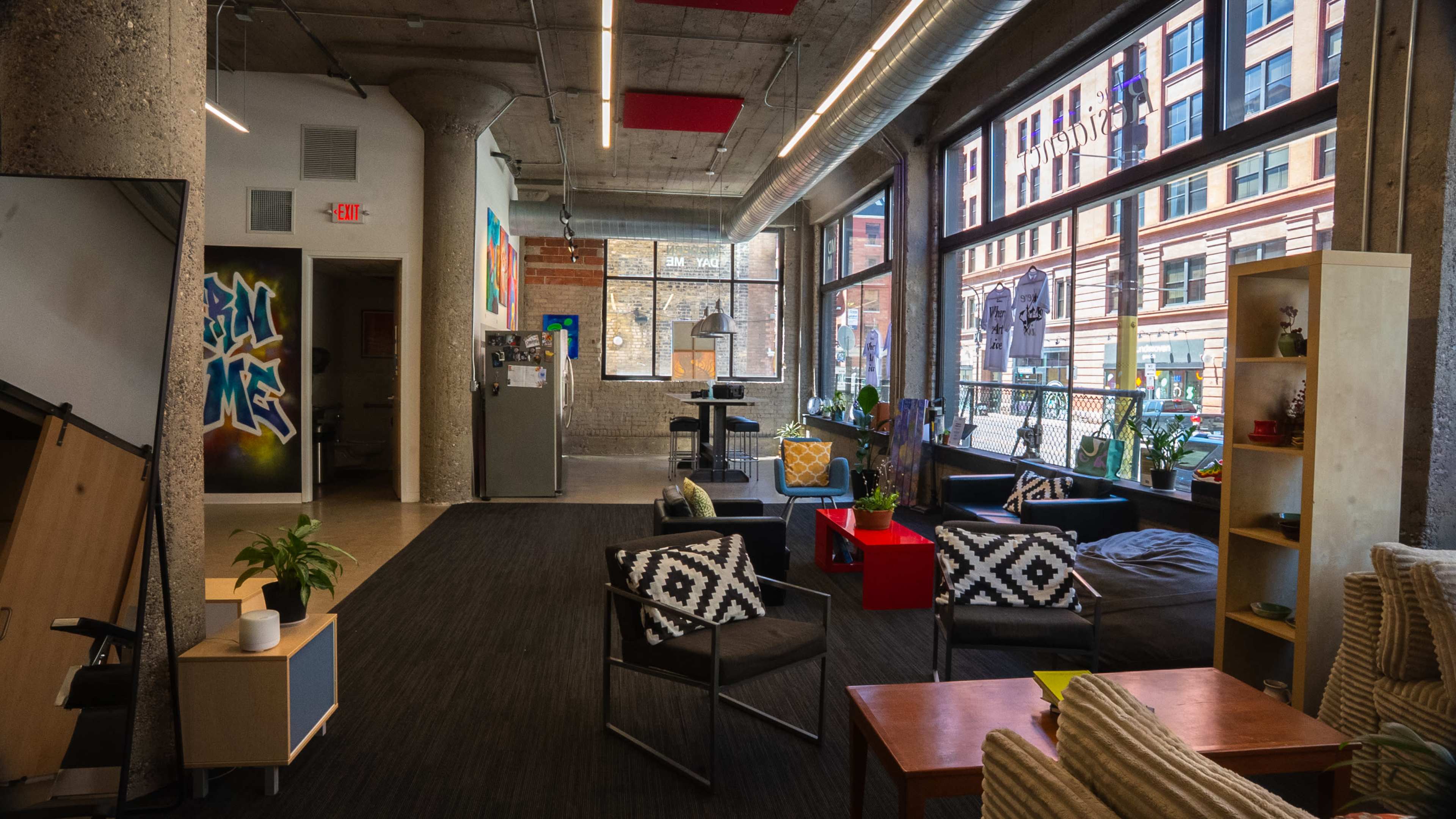 The image shows an open office space with large windows, modern furniture, and a variety of plants, featuring areas for seating and collaboration.