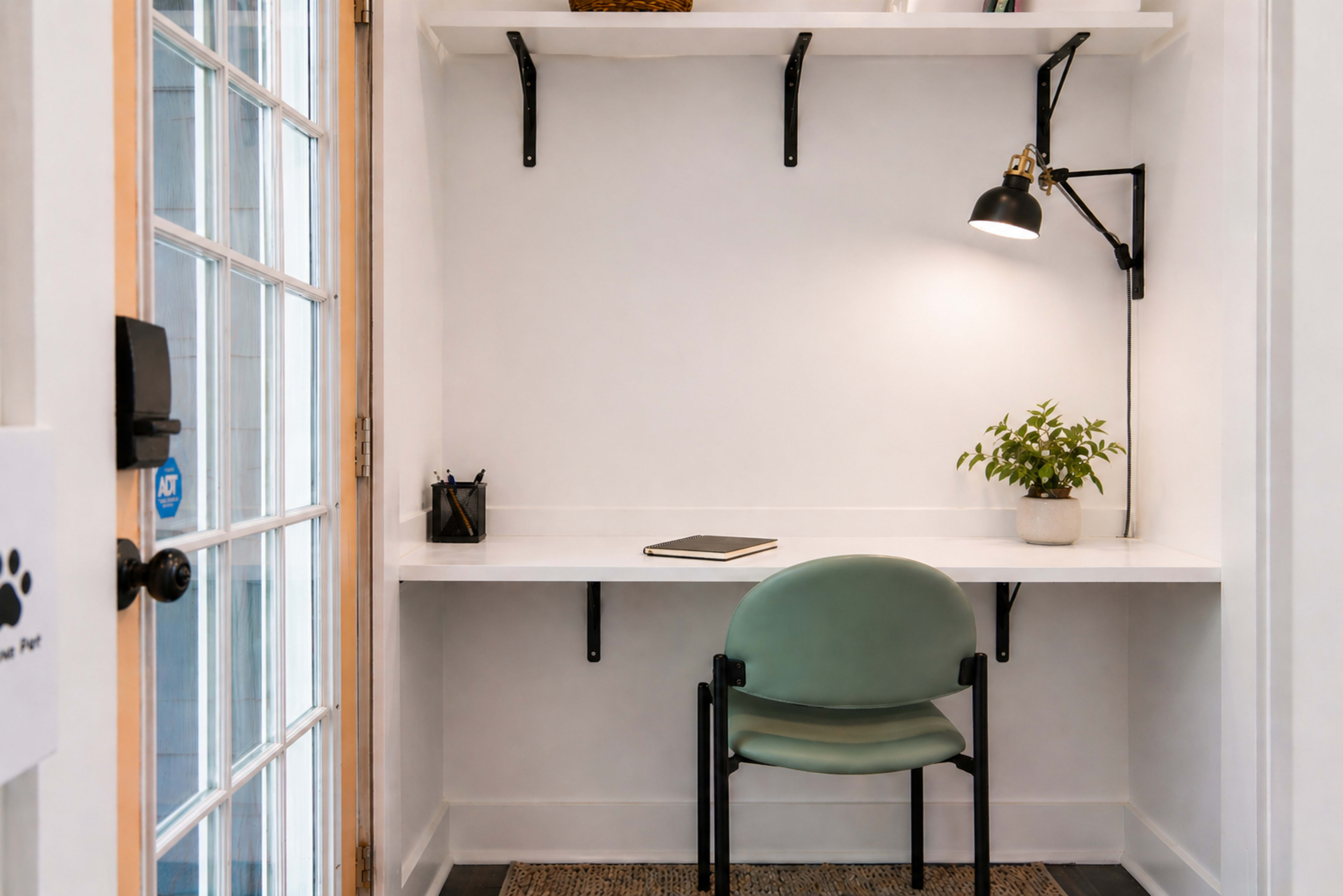 The image shows a small, minimalist workspace featuring a wall-mounted desk, a green chair, a potted plant, and a wall lamp.