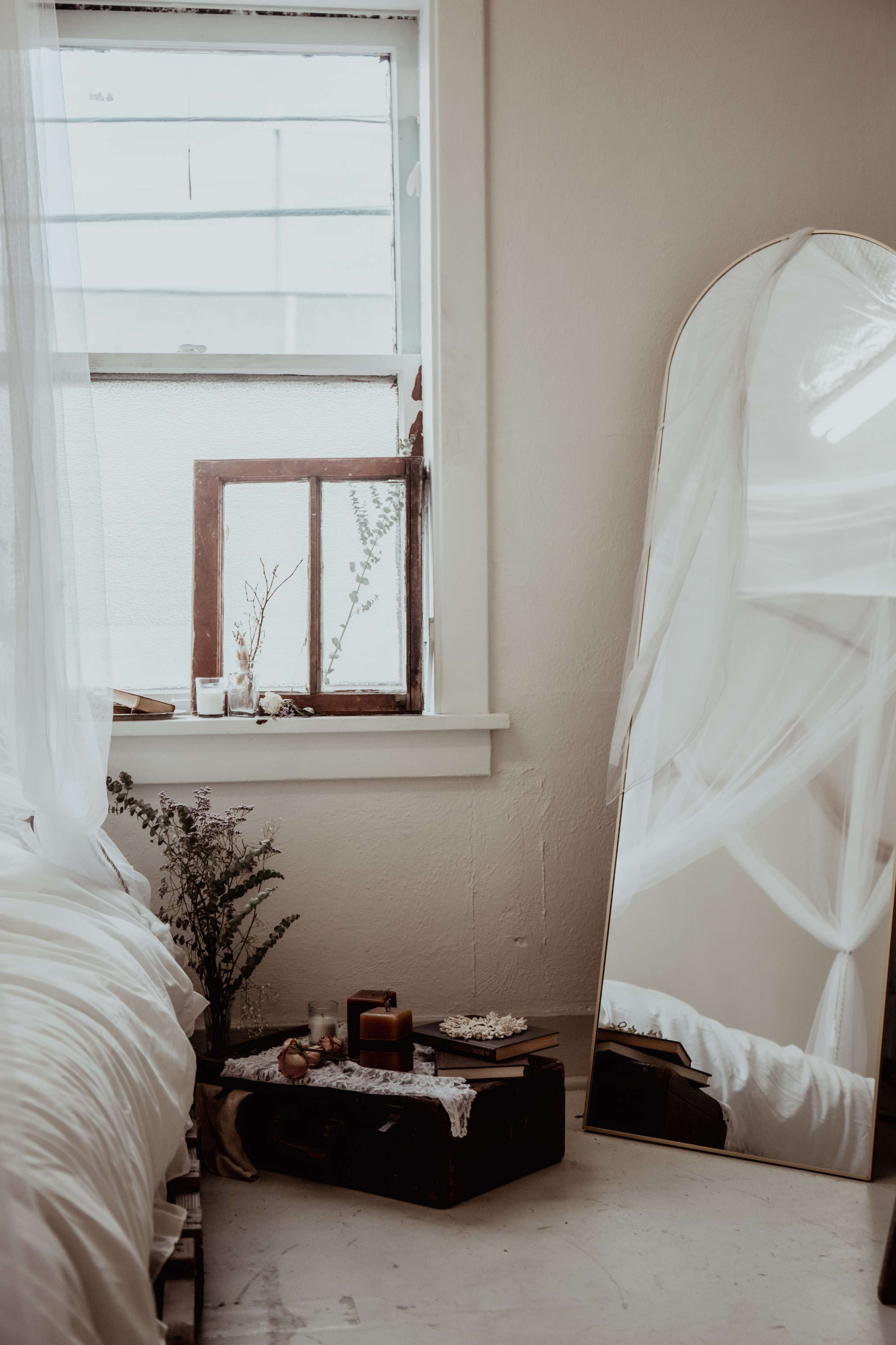 A corner of a bedroom featuring a bed with white bedding, a large mirror, a windowsill with plants, and a small table with decorative items.