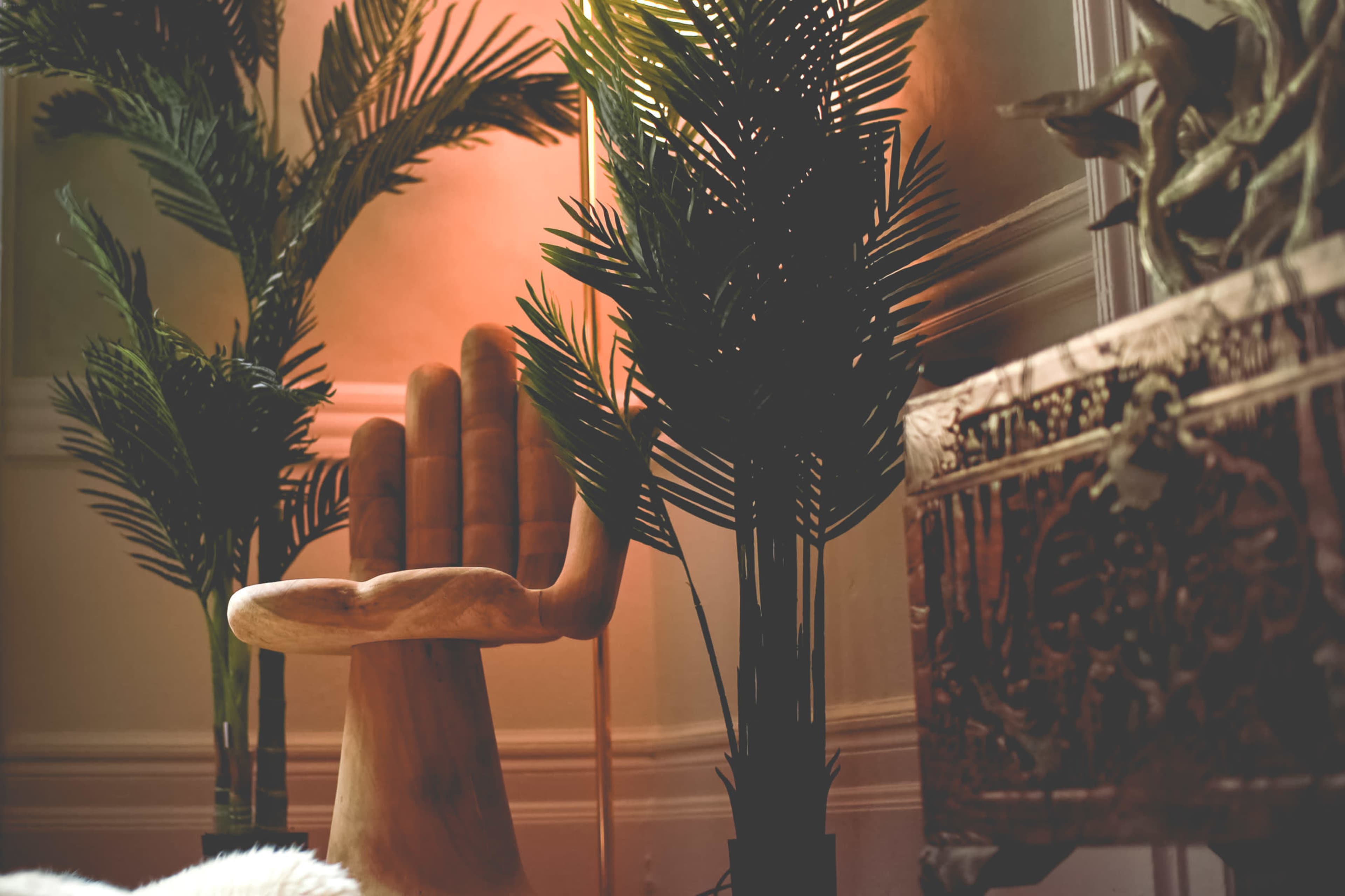The image features a wooden hand-shaped chair surrounded by potted palm plants and an intricately designed decorative box.