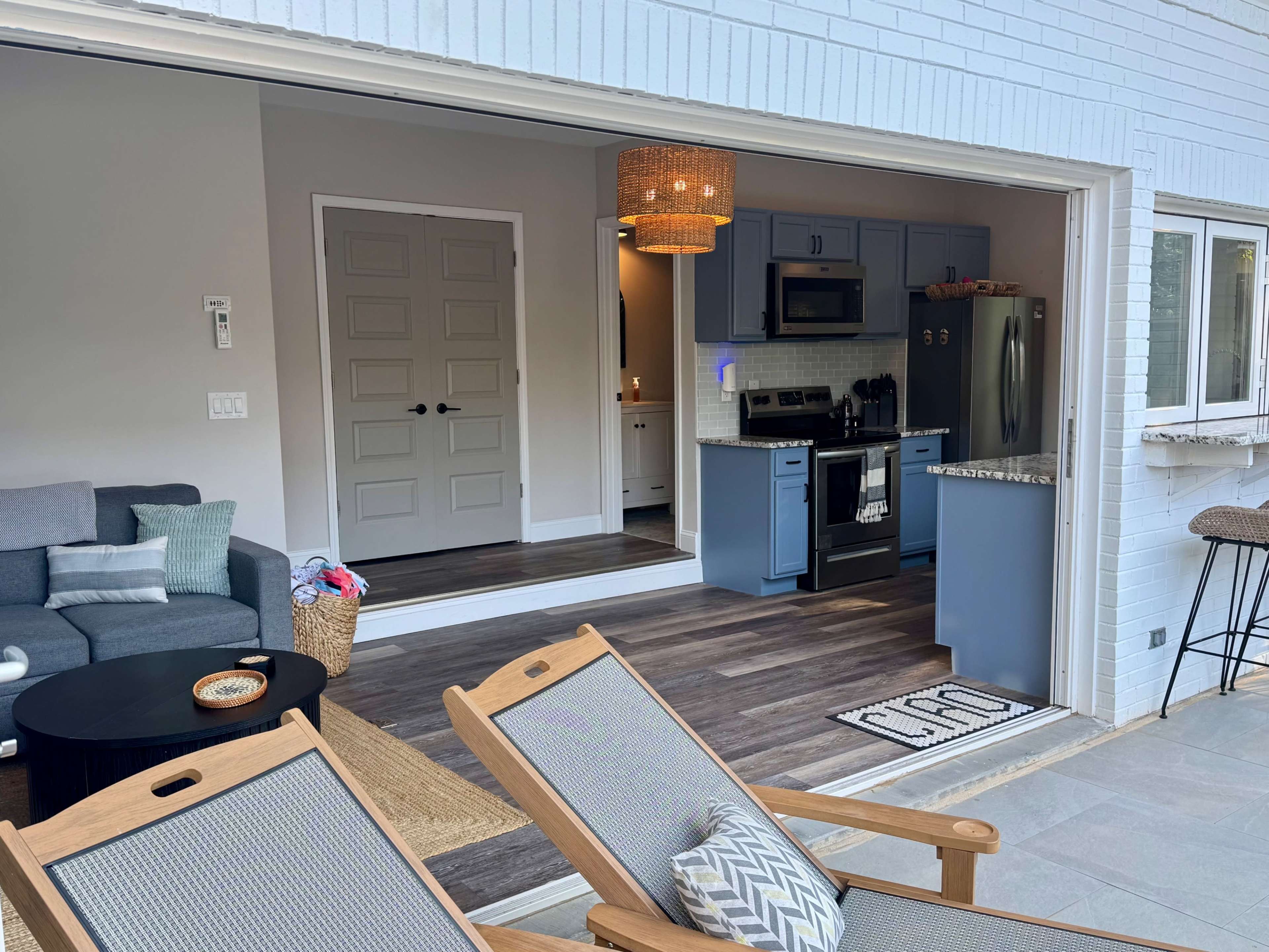 The image shows a bright, open-concept living space featuring a kitchen with blue cabinets and an adjacent lounge area with outdoor seating.