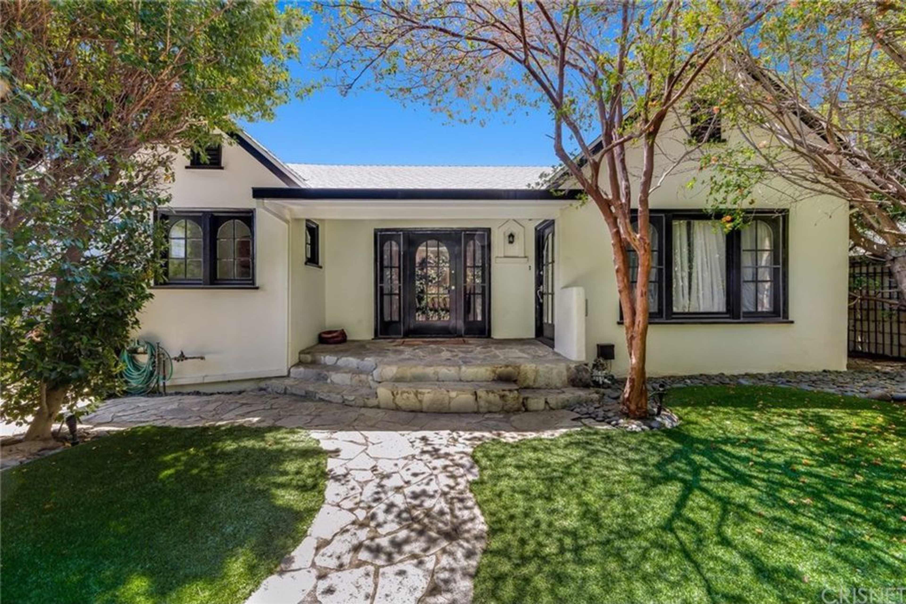 The image shows a charming white house with a stone pathway leading to a front entrance, surrounded by green grass and trees.