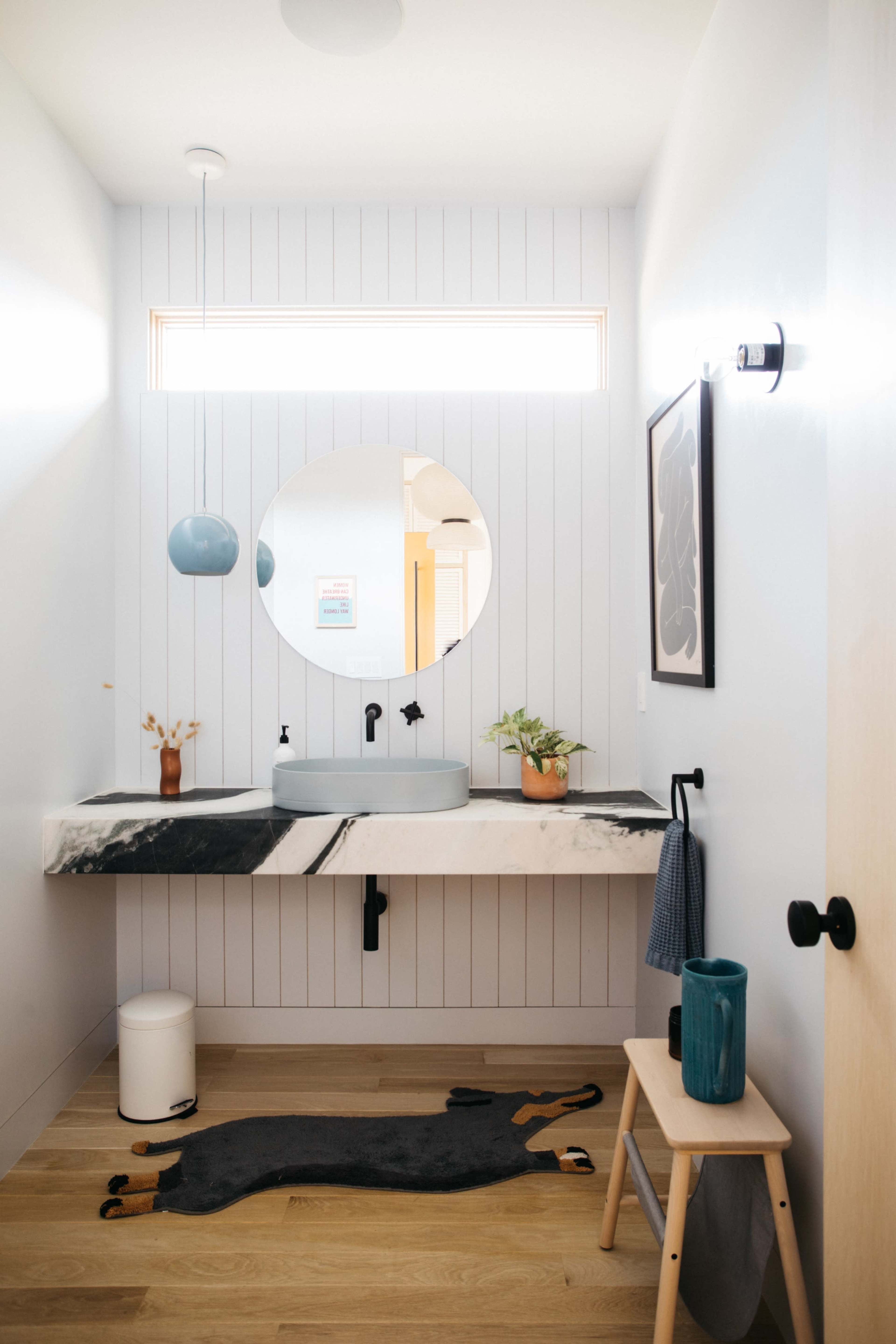 A modern bathroom features a round mirror above a minimalist sink with a floating countertop, wood flooring, and a small plant beside a decorative stool.