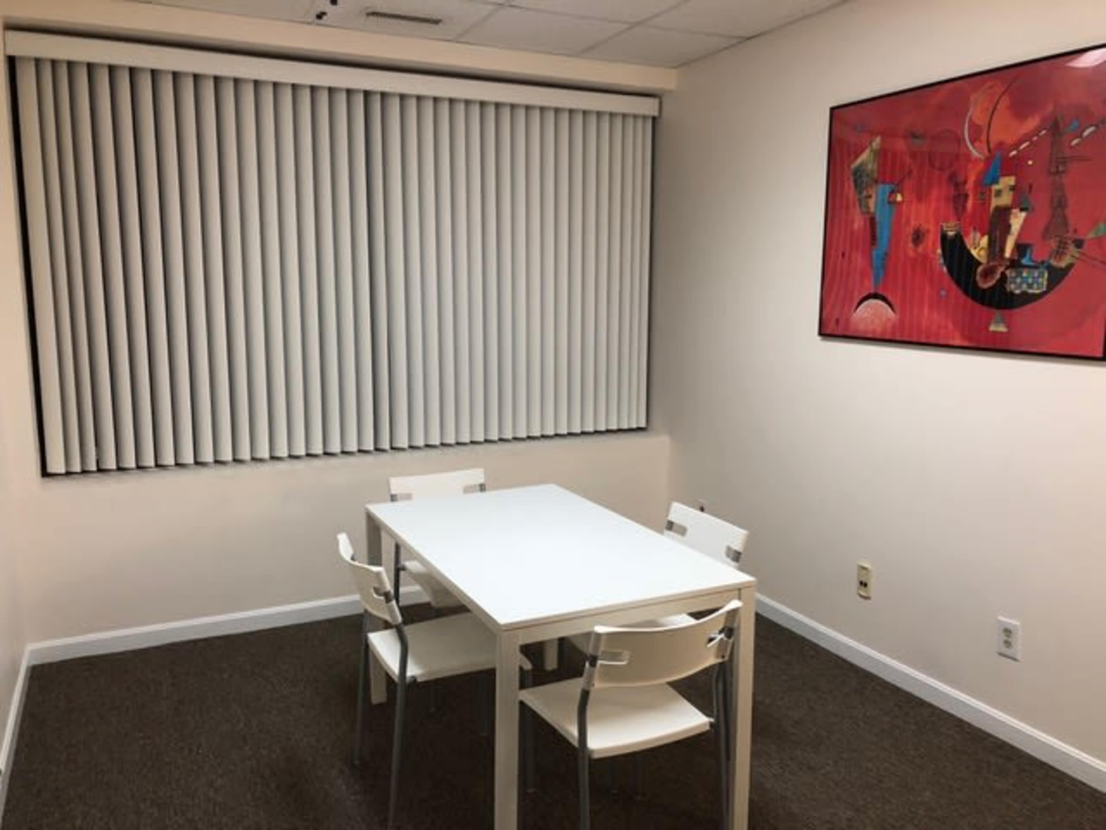 A small room features a white table surrounded by four white chairs, with vertical blinds over a window and a framed artwork on the wall.