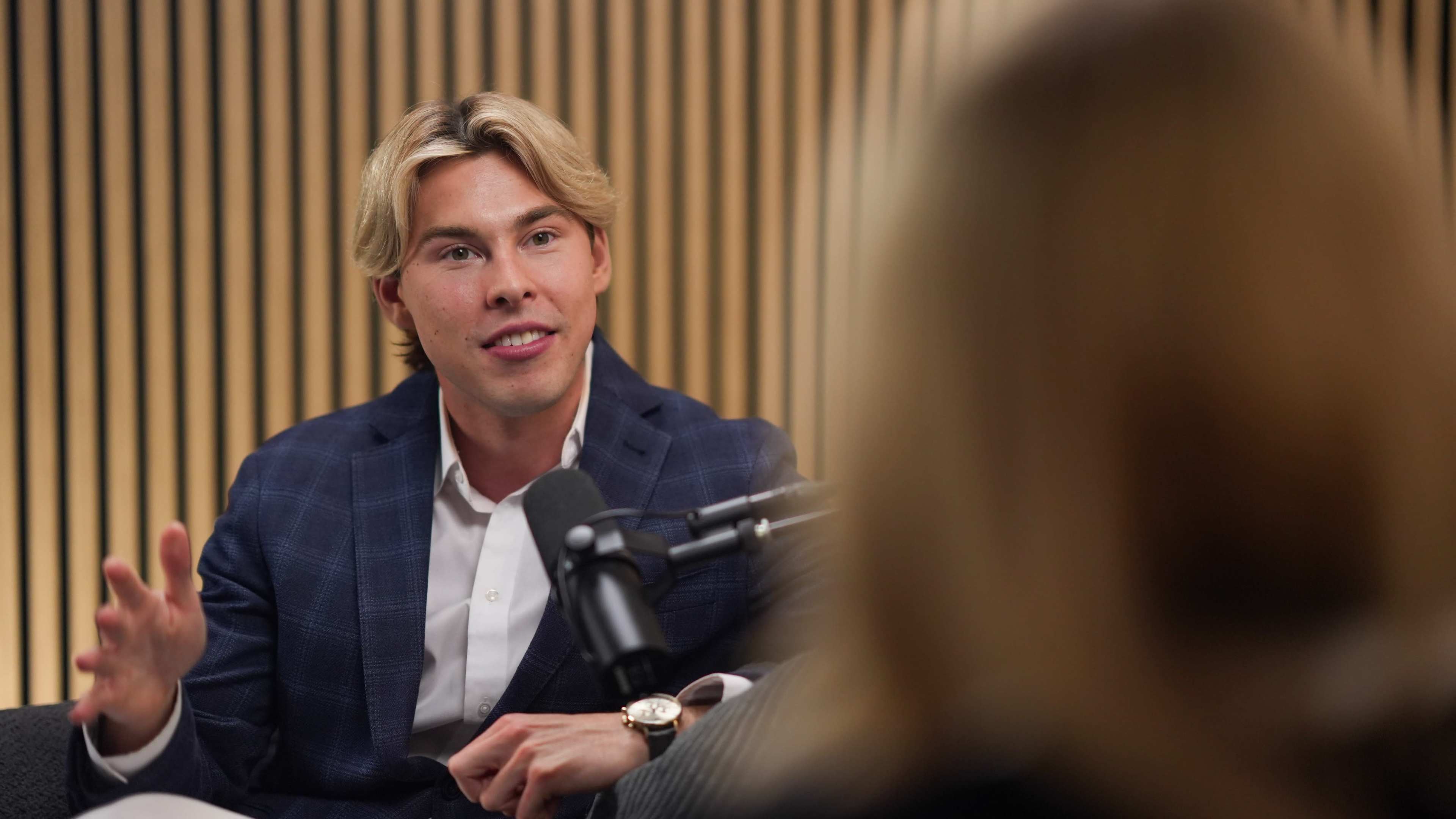 A person in a suit is speaking into a microphone during a podcast interview, with another individual partially visible in the foreground.