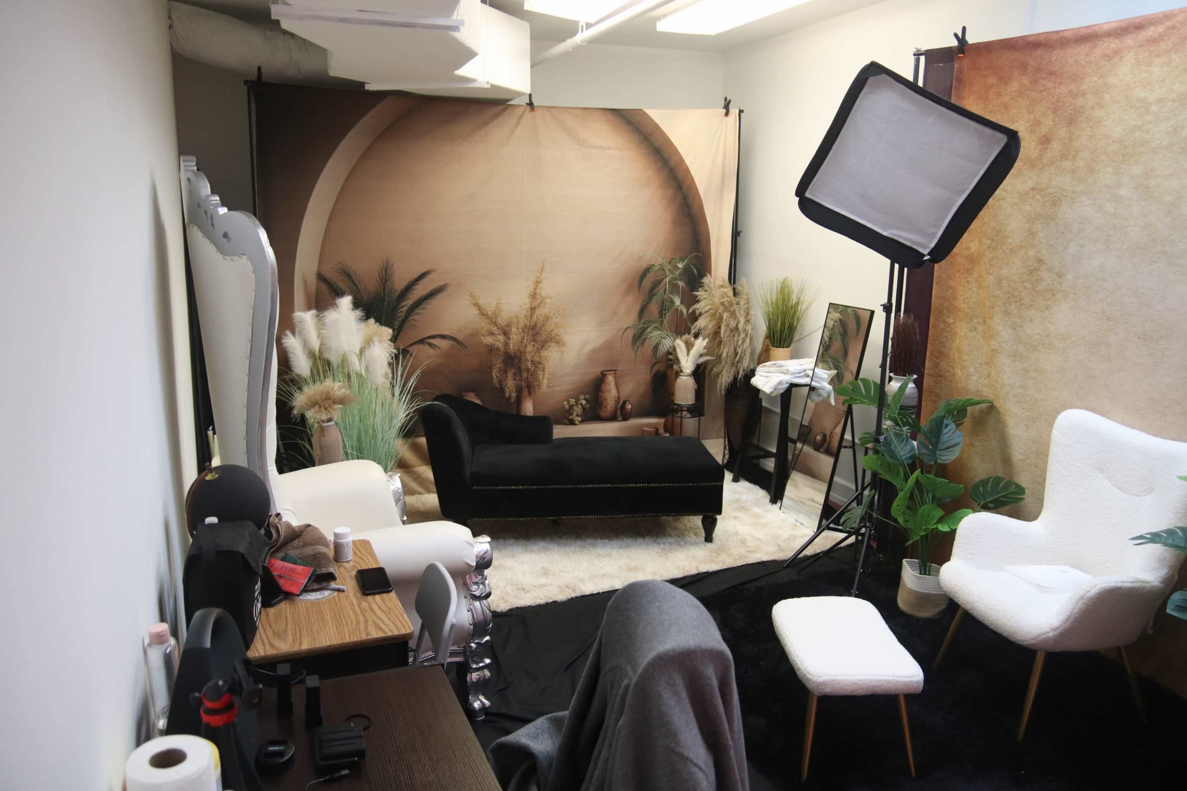 The image shows a photography studio with a styled set featuring a black sofa, decorative plants, and a backdrop of neutral tones.