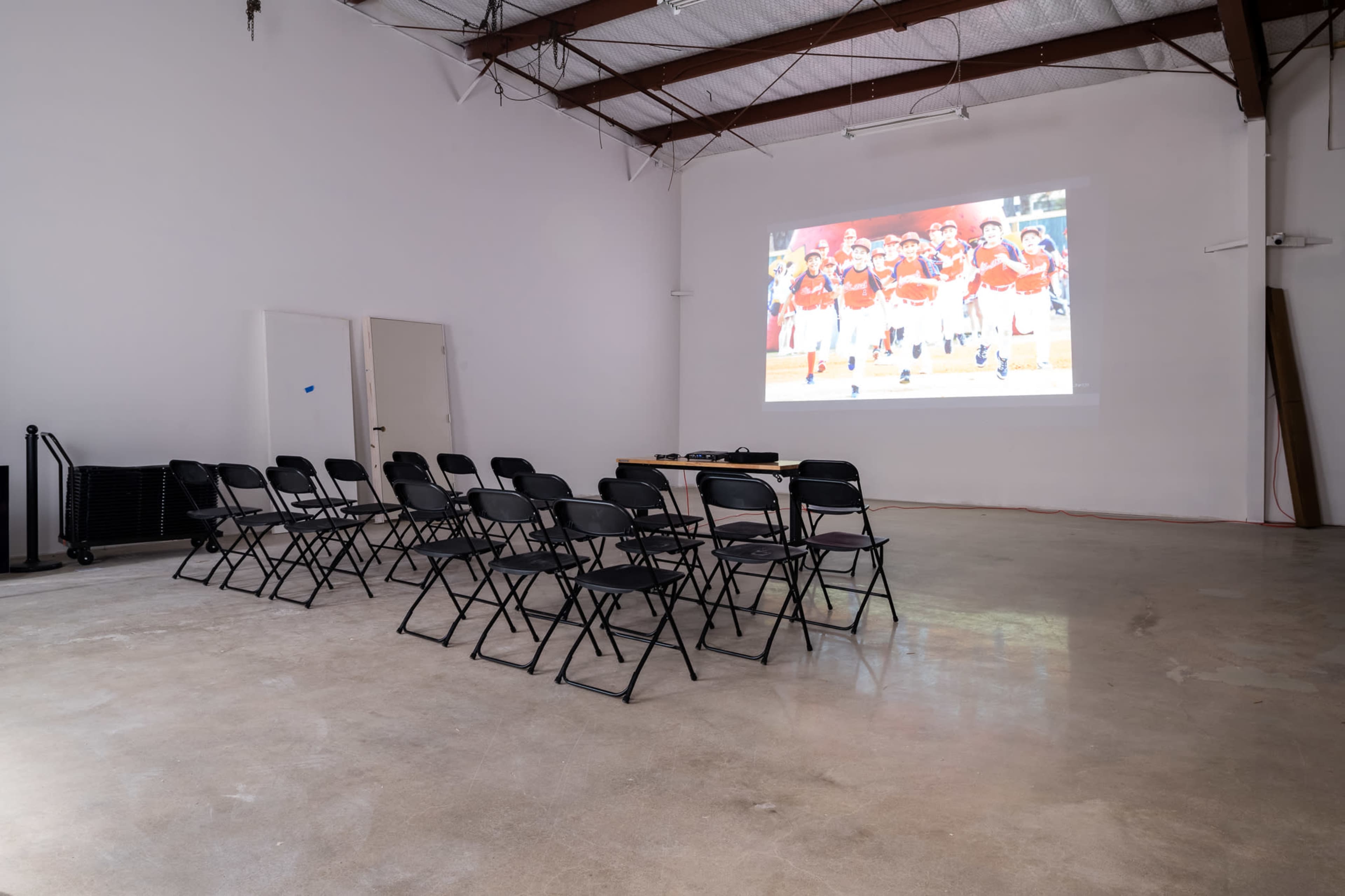 A row of black folding chairs faces a projector screen displaying an image of people in uniforms, set in a spacious, empty room with concrete flooring.