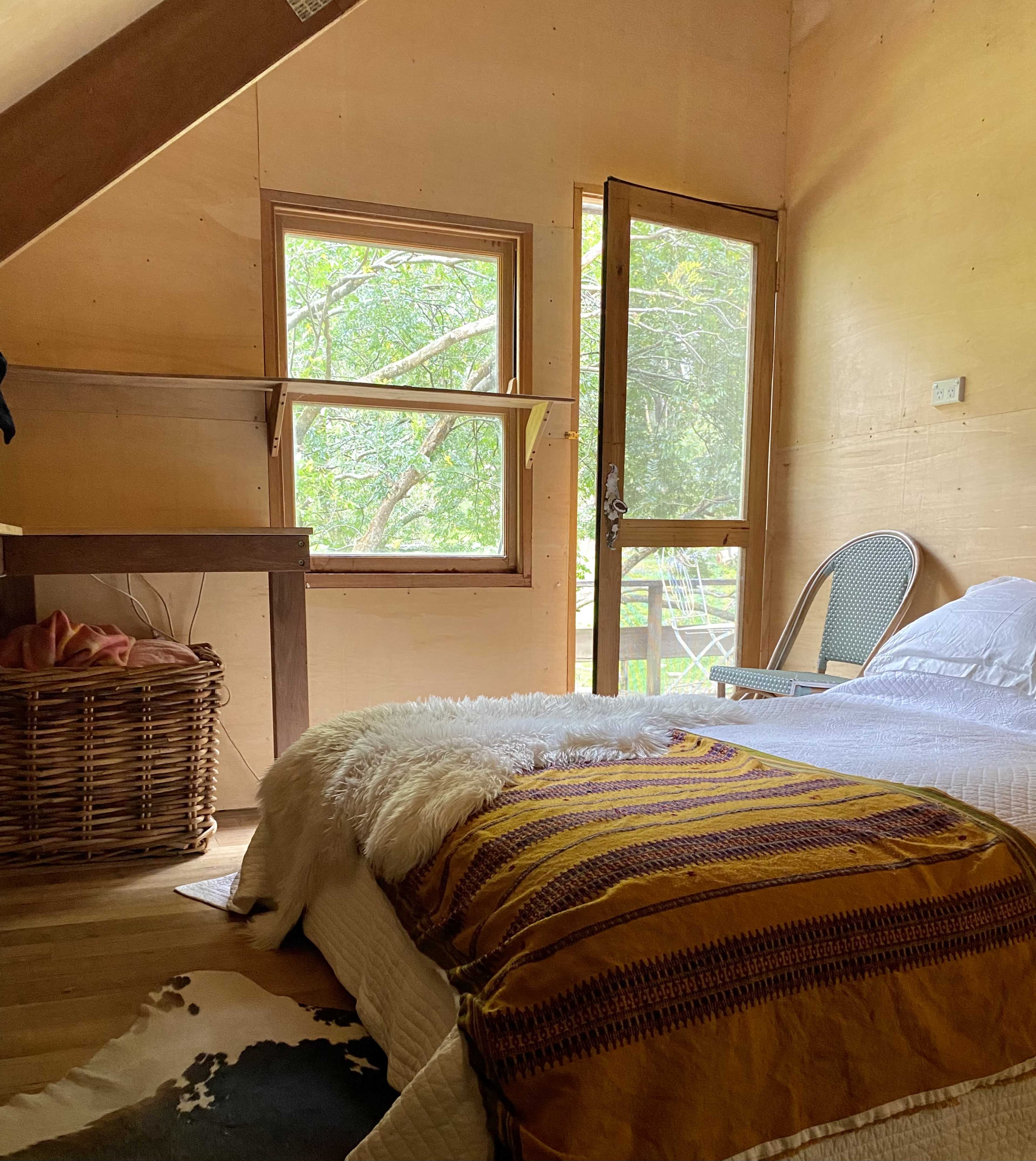 A cozy bedroom with a bed, a woven basket, and a window open to a view of greenery outside.