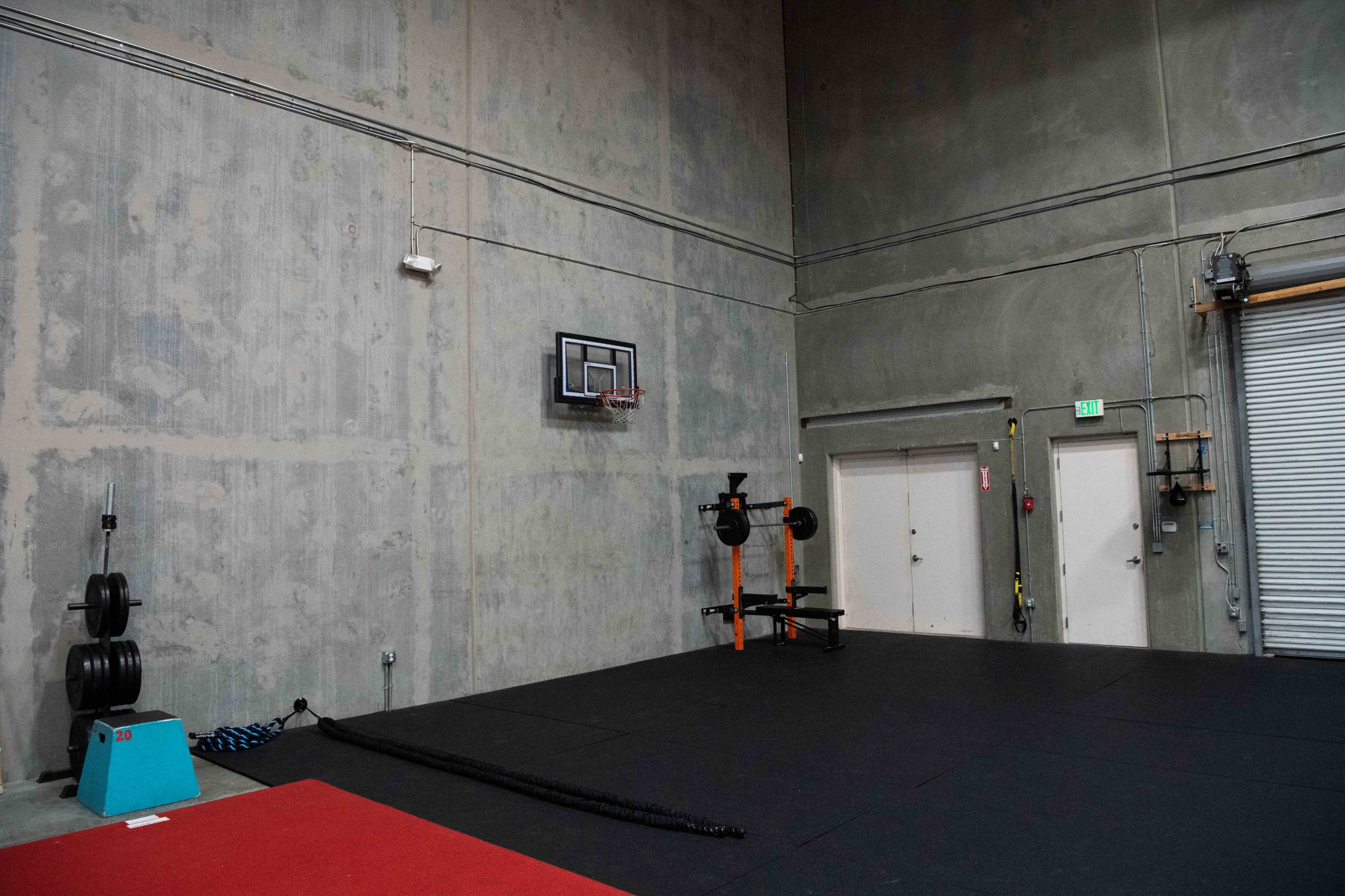 A concrete gym interior with a basketball hoop mounted on the wall, a barbell set on a rack, and a section of rubber flooring.
