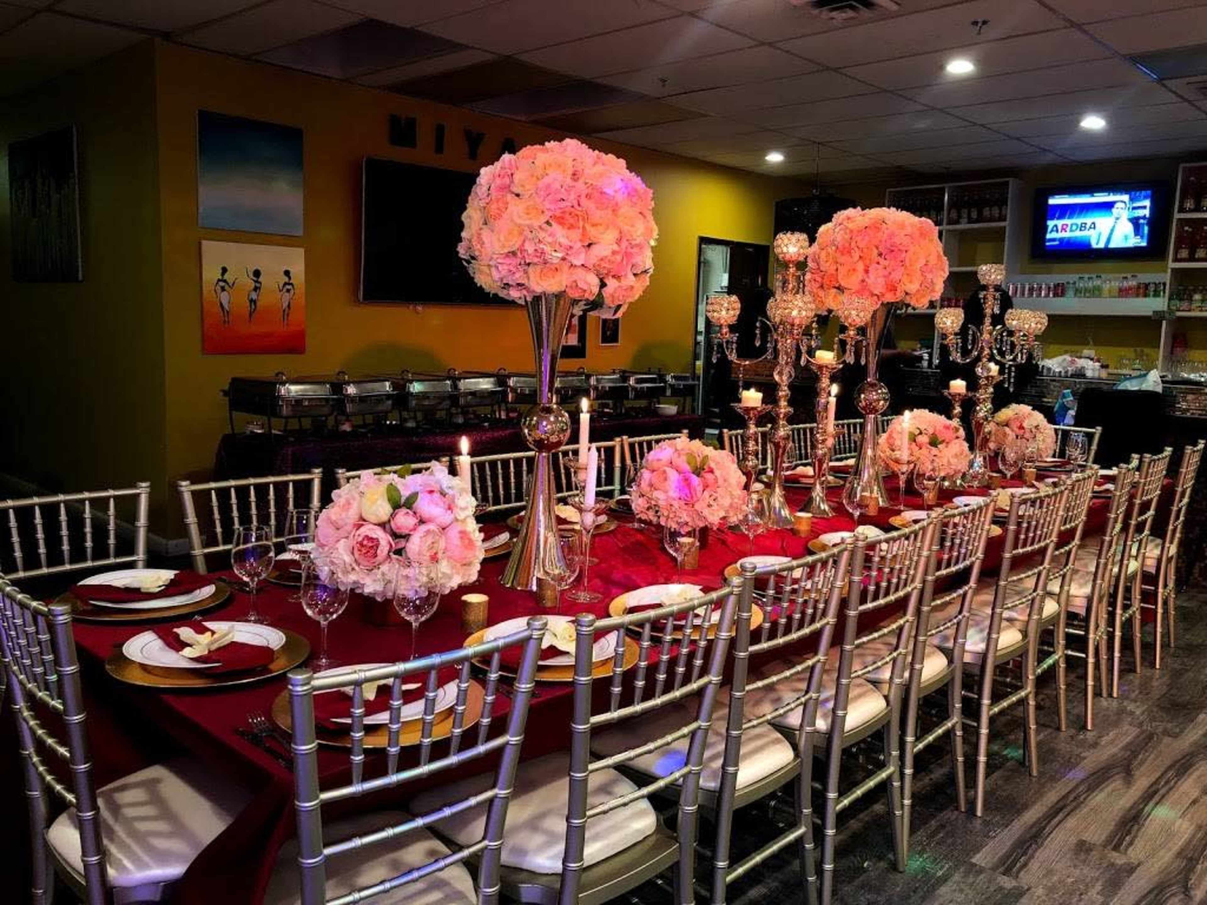 A lavishly set dining table with elegant centerpieces of pink flowers and candles, surrounded by gold chiavari chairs in a brightly lit restaurant.