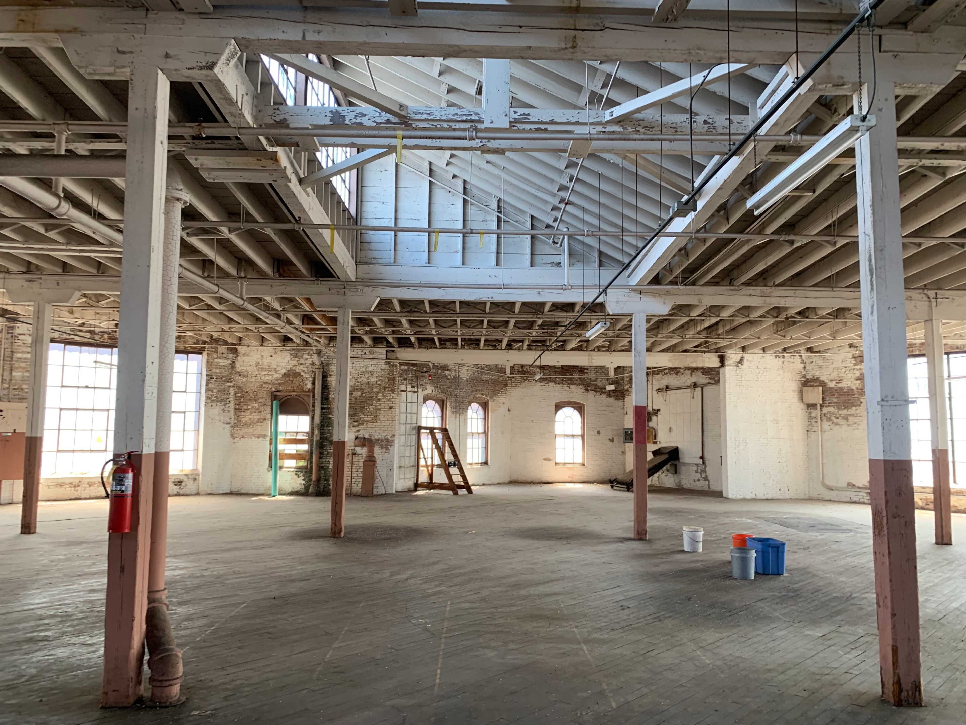 The image shows an empty industrial space with high ceilings, exposed beams, and several large windows.