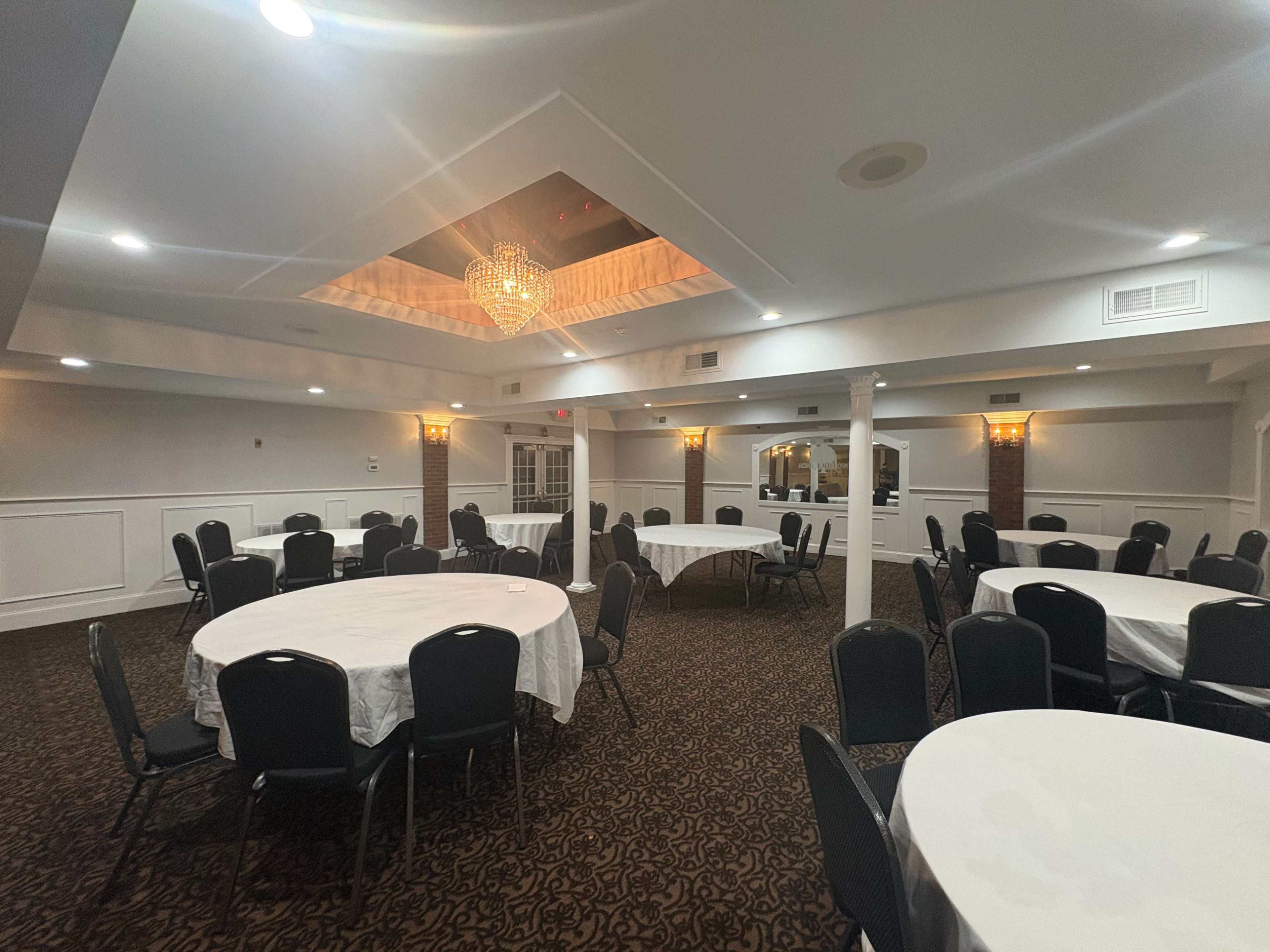 The image shows a neatly arranged banquet hall with round tables covered in white tablecloths and black chairs, featuring a chandelier and decorative ceiling.
