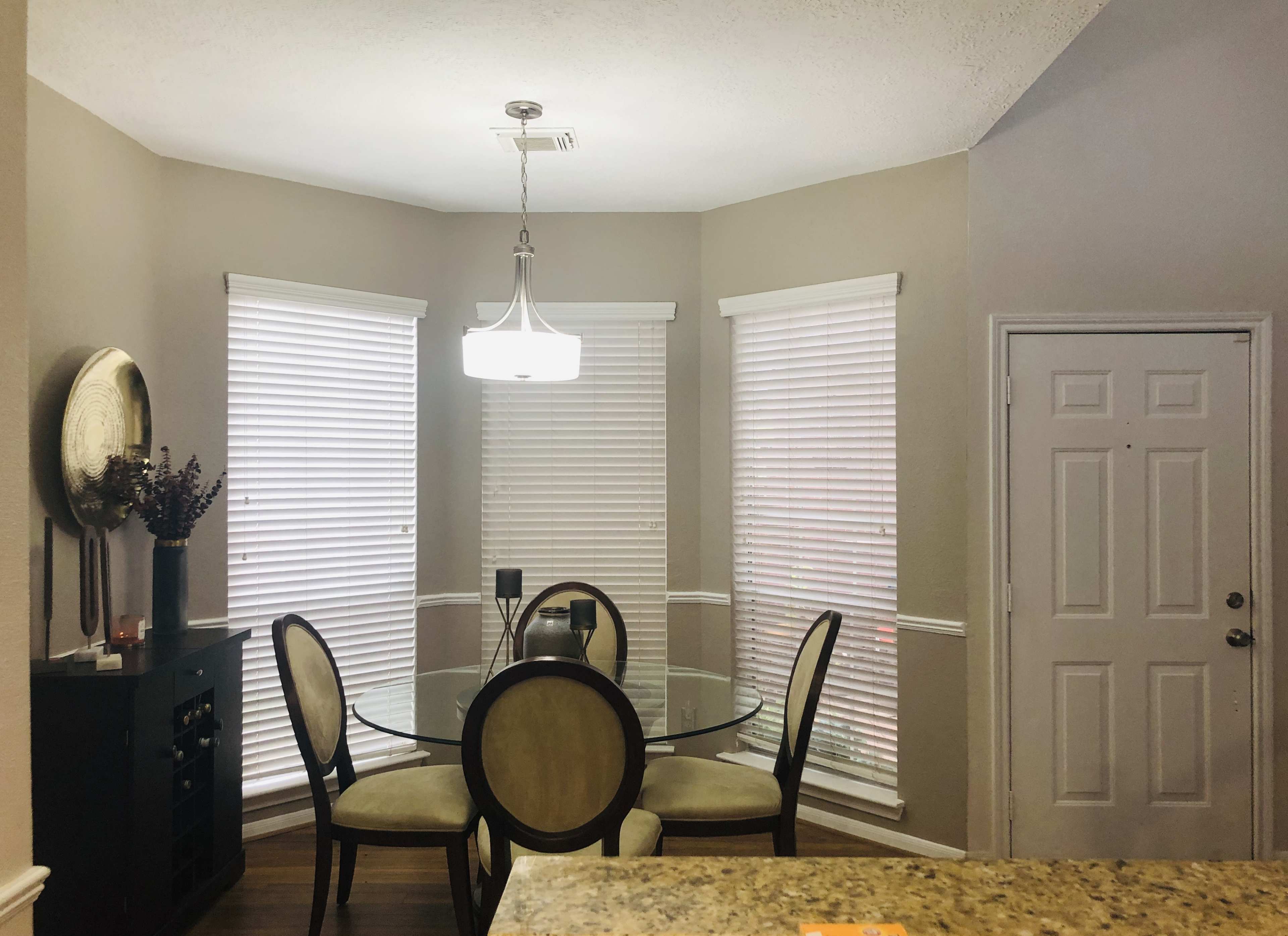 A dining area features a round glass table surrounded by four chairs, with a pendant light overhead and large windows with blinds.