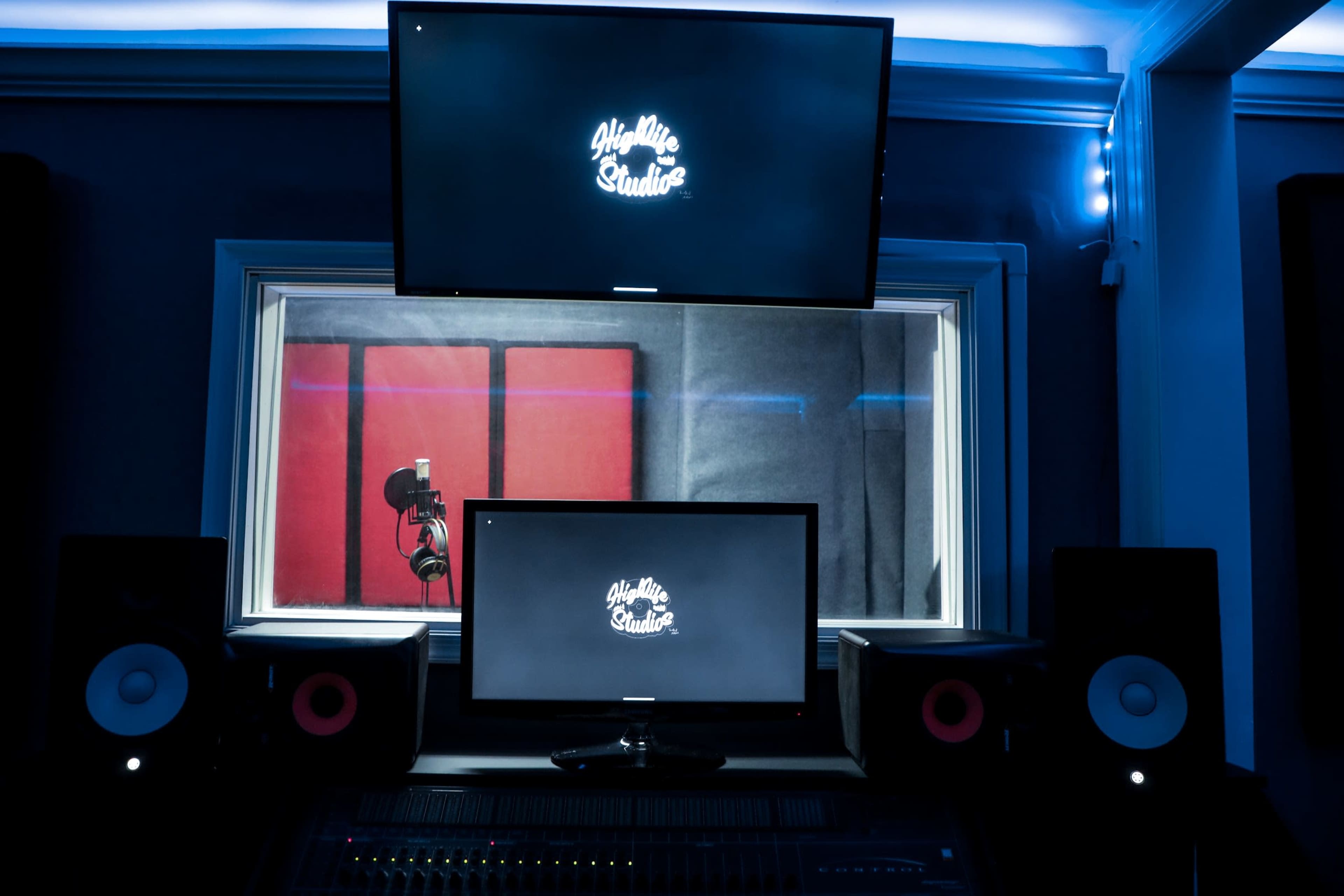 A sound recording studio with two monitors displaying the logo "You Mic Studio," a microphone in a soundproof booth, and studio speakers on either side of a mixing console.
