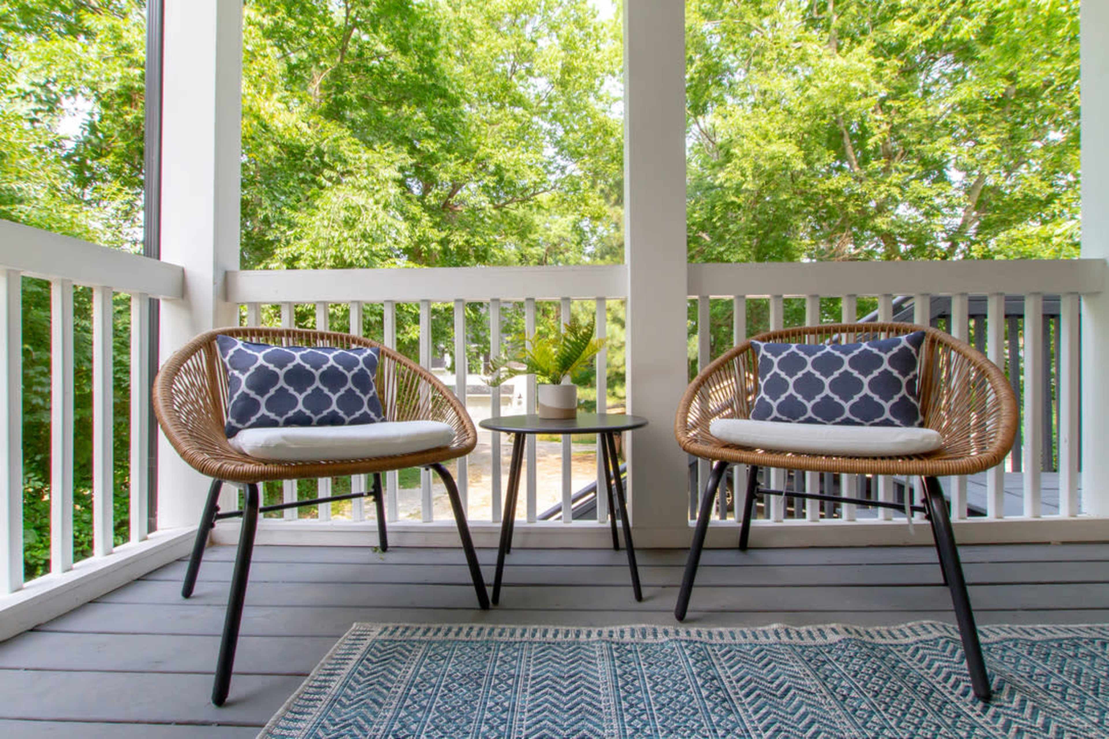 The image shows a porch with two wicker chairs adorned with patterned cushions, a small table between them, and greenery in the background.