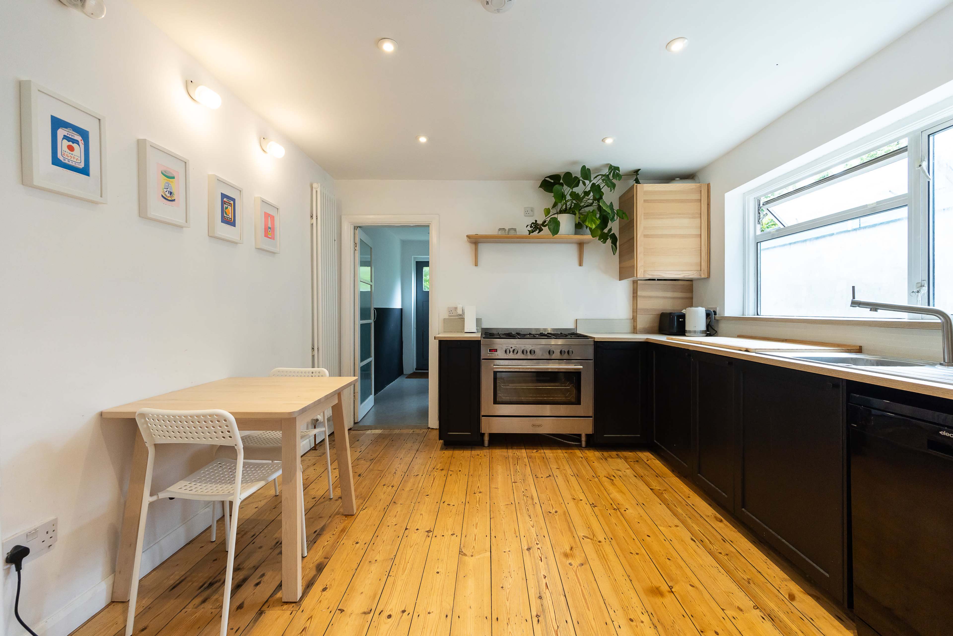 The kitchen features a wooden floor, stainless steel appliances, a wall-mounted shelf with potted plants, and a small dining table with two chairs.
