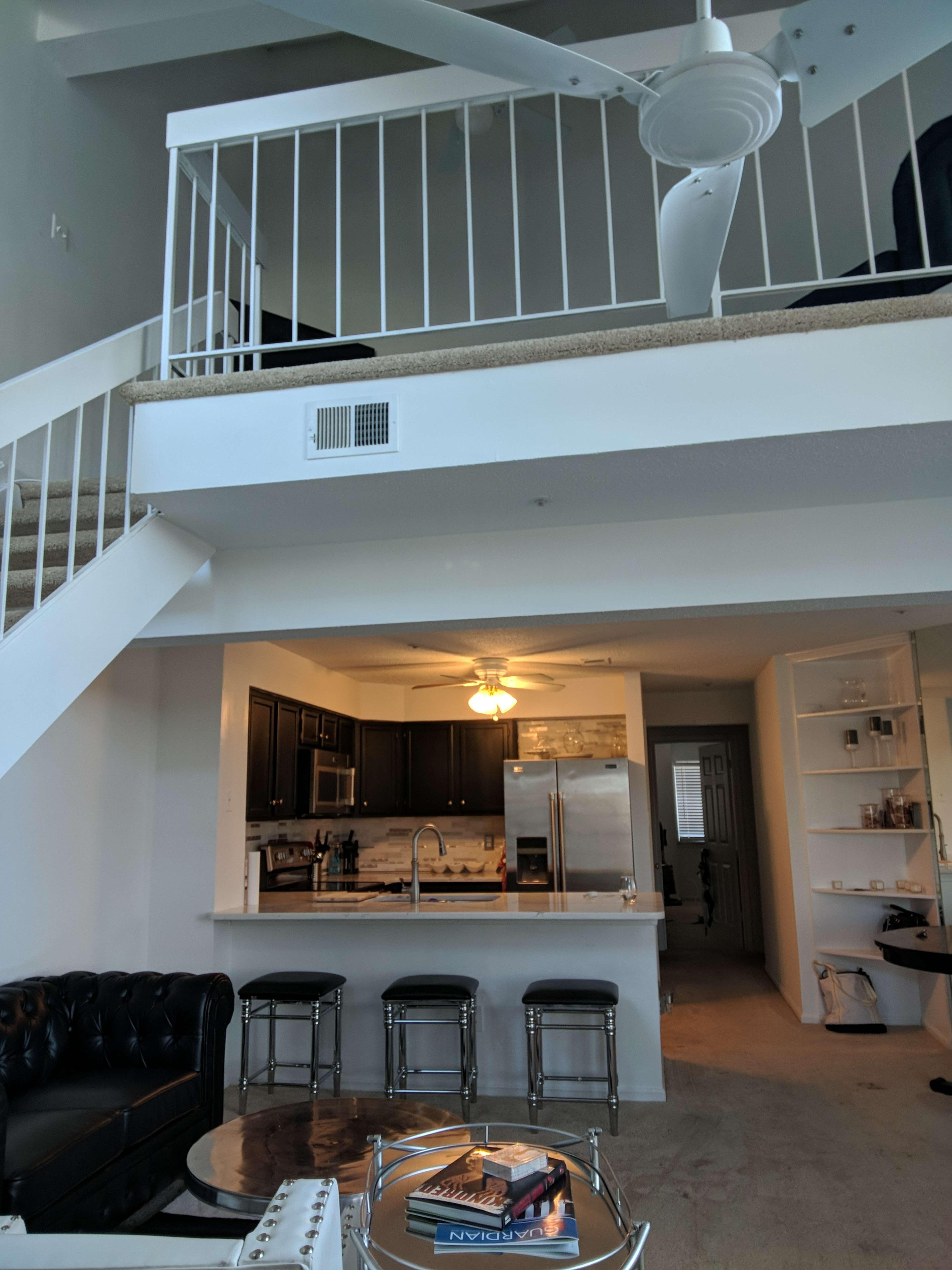 A modern living space featuring a kitchen with dark cabinets, a living area with a black leather sofa, and a lofted bedroom area above.